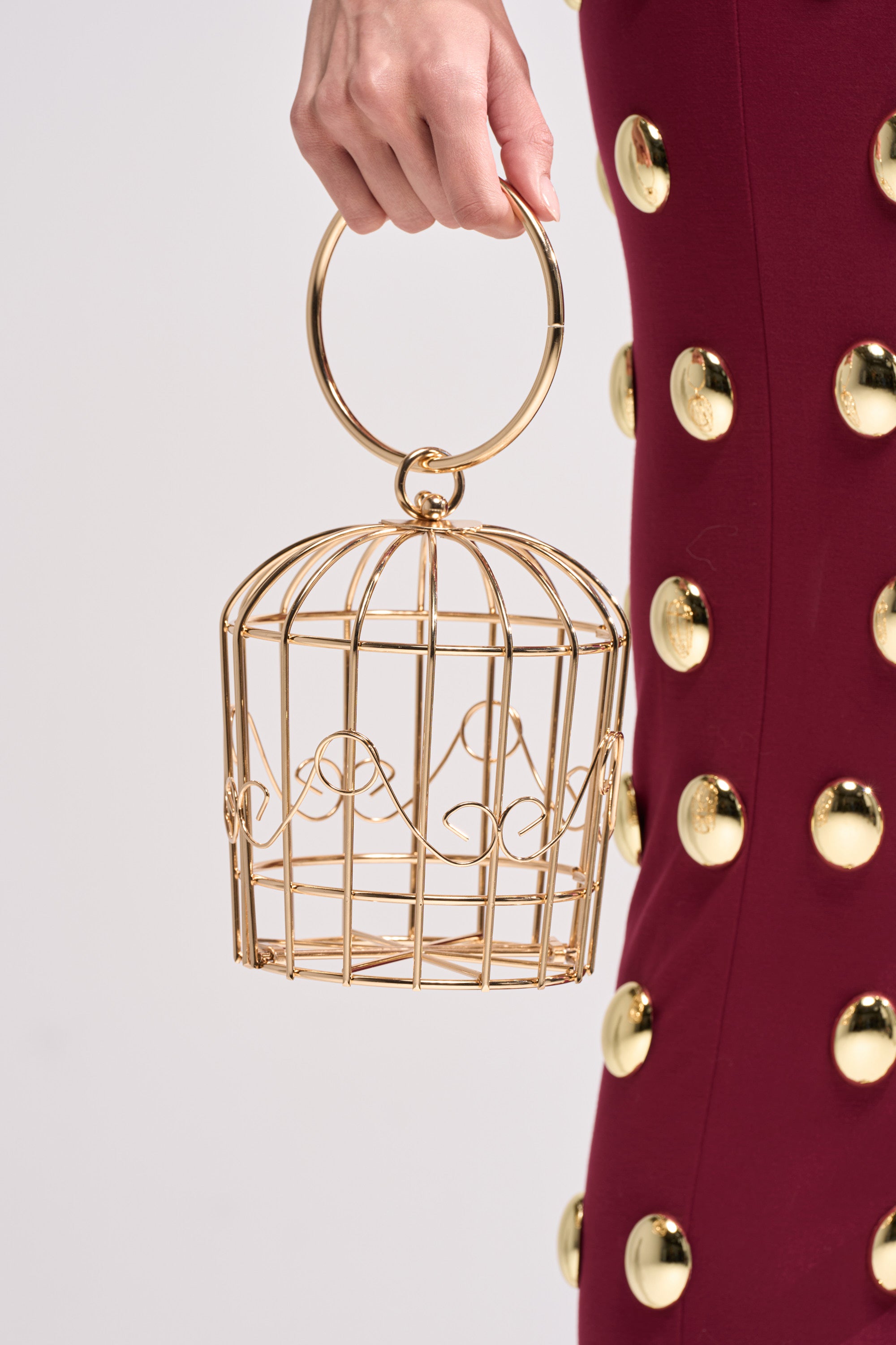 A person holds the NEVER CAGED PURSE by its round handle, pairing it with a burgundy skirt featuring large gold buttons.