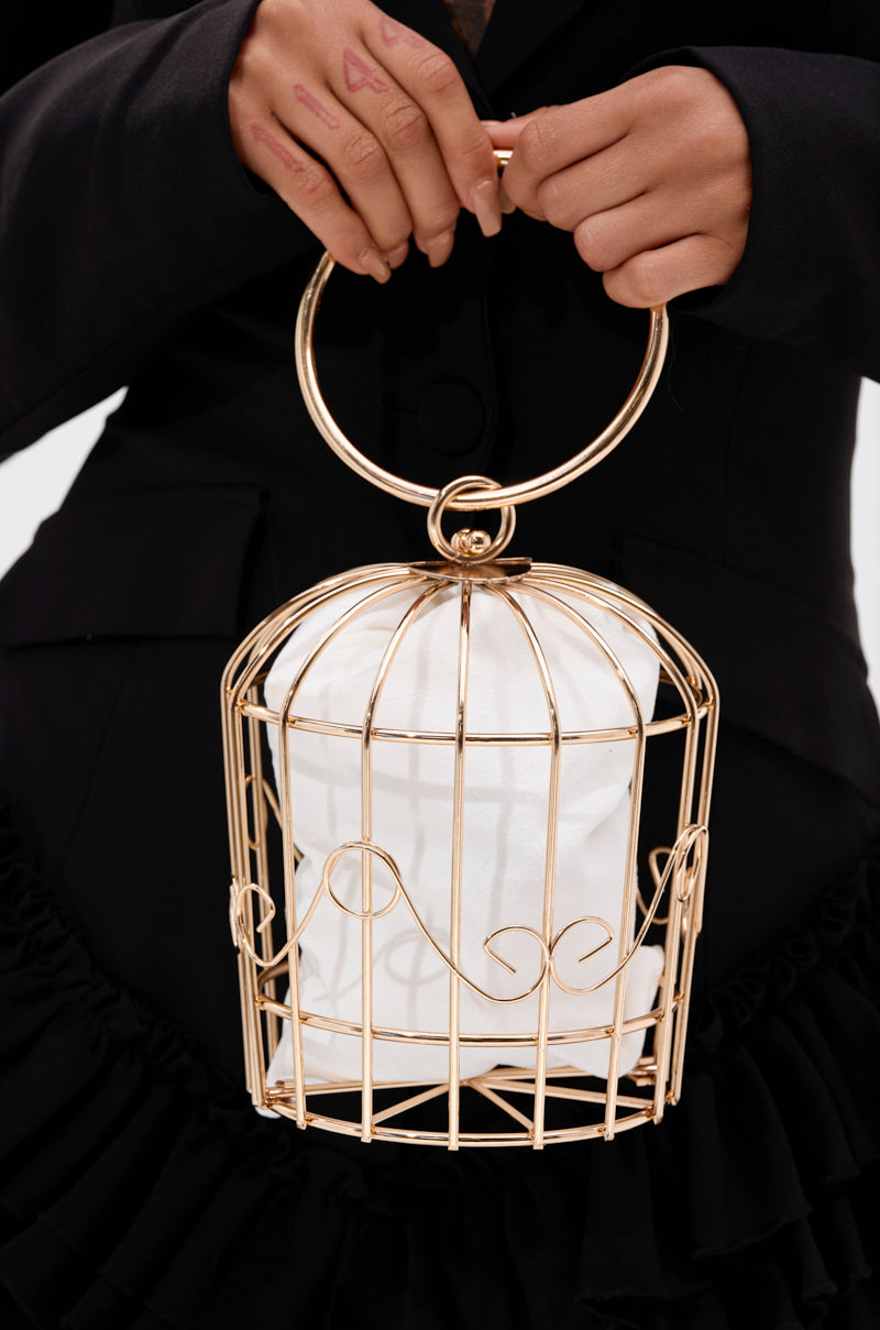 A person in black holds the NEVER CAGED PURSE, featuring a gold cage design with a large round handle and a white fabric pouch inside. A small heart mark on their hand completes this sculptural statement look.