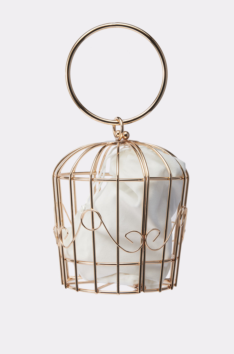 The NEVER CAGED PURSE features a gold wire birdcage design, a large circular handle, and a white cloth pouch inside—this sculptural statement bag stands out against any plain background and is sure to turn heads.