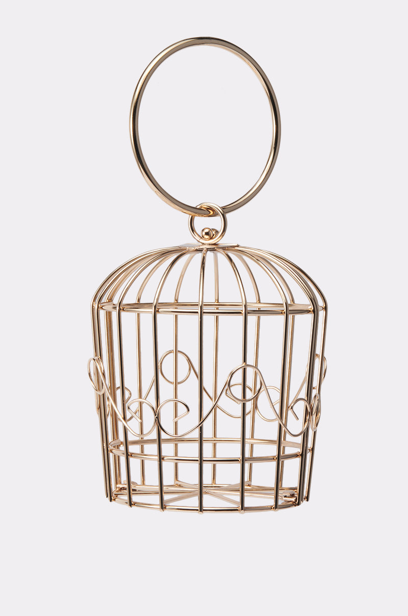 The NEVER CAGED PURSE is a small, ornate gold-colored wire statement bag with decorative curved detailing and a large circular top handle, displayed empty against a plain white background. 4