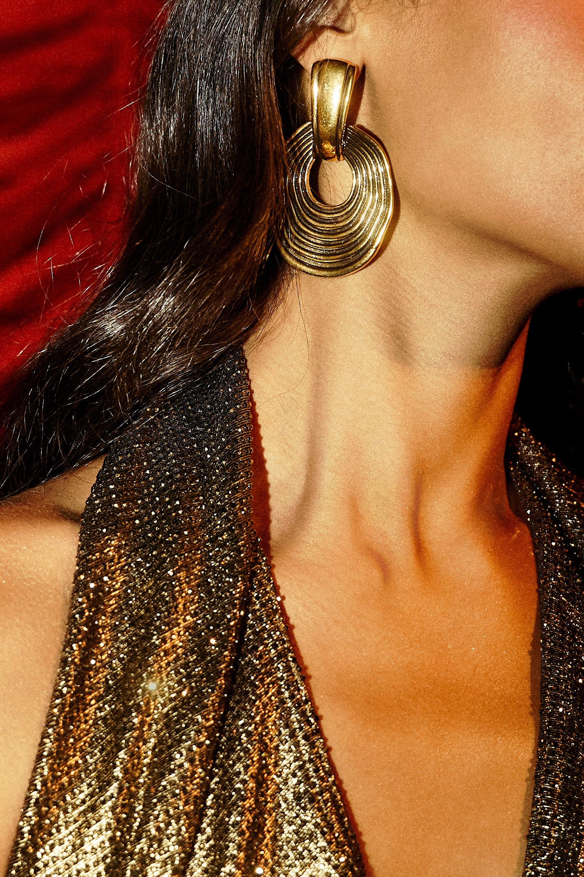 A close-up of a woman in a sparkly gold dress, wearing the SO WAVY EARRING with her wavy dark hair draped over her shoulder, set against a red background. 1