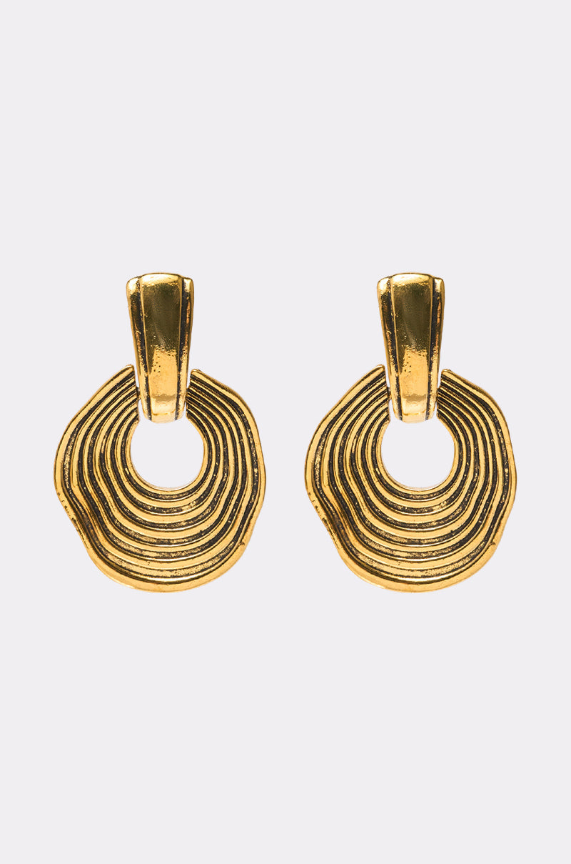 The SO WAVY EARRING features gold drop earrings with textured, concentric circular designs and open centers on simple gold studs, creating a striking statement look against a light gray background. 0