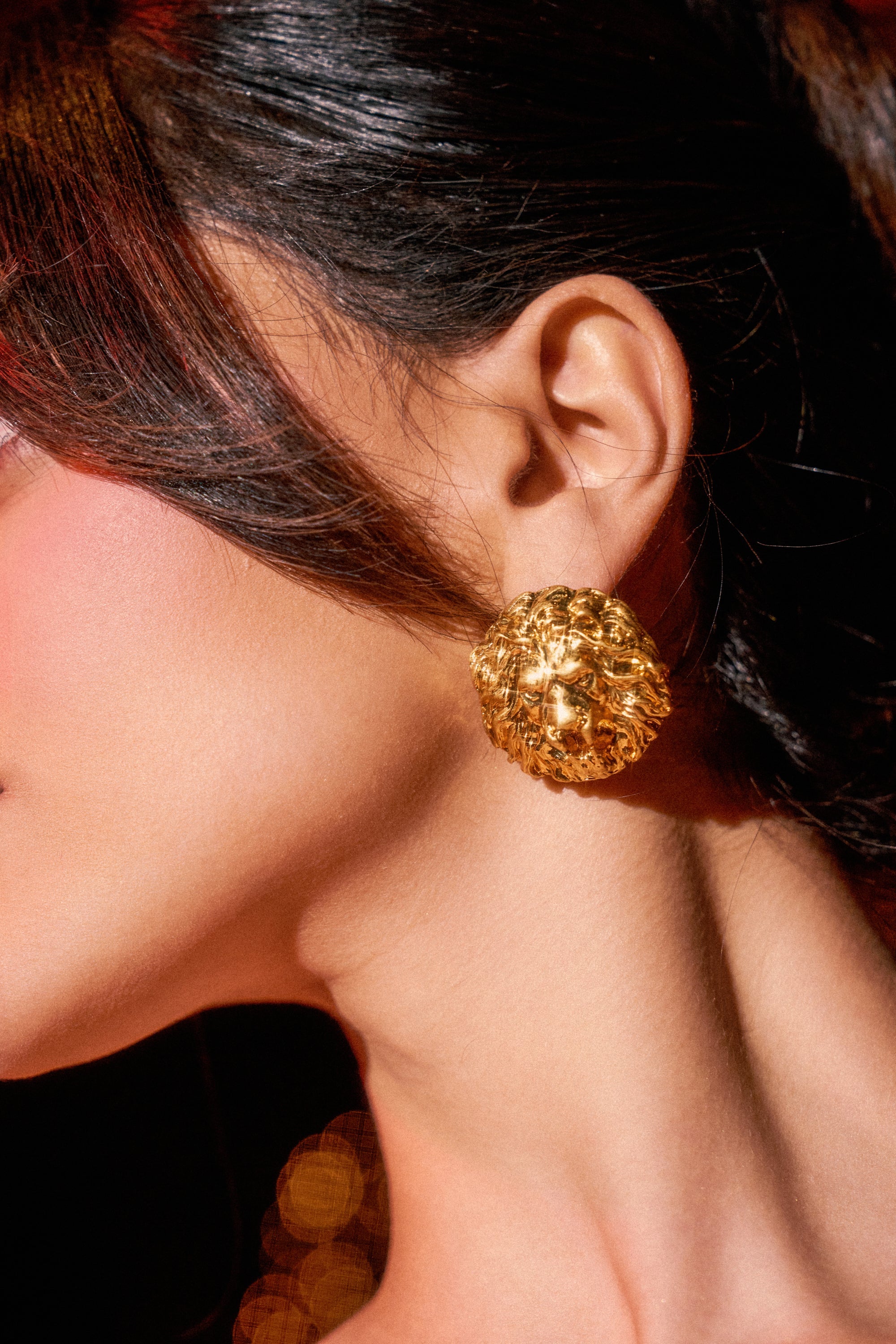 A woman's ear is adorned with the ROAR EARRING—a large, textured, round gold statement piece. Her dark hair is swept back to reveal smooth skin and a soft blush, set against a dark, blurred background. 0