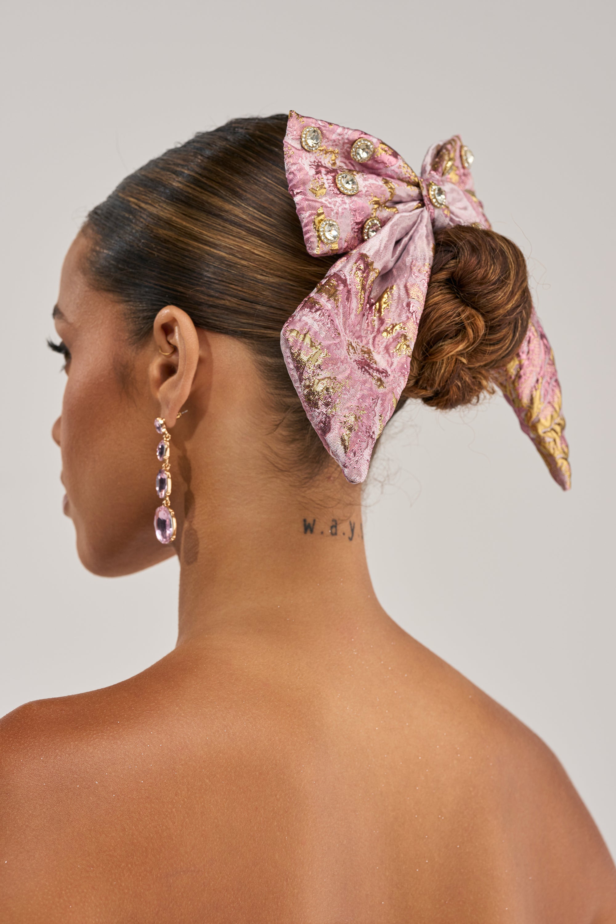 A woman seen from behind with her hair in a bun, styled with the GLAM GROOVE BROCADE HAIR BOW featuring pink and gold fabric and gems. She wears dangling earrings and has a “w.a.y” tattoo on her neck.