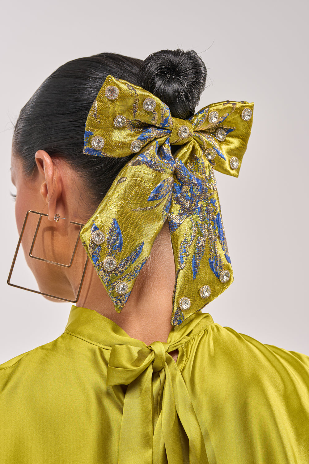 A woman with dark hair in a sleek bun wears the LETS GO EMBELLISHED HAIR BOW, featuring blue patterns and rhinestone details. She pairs it with a matching satin top and oversized square gold earrings, shown from the back. 0