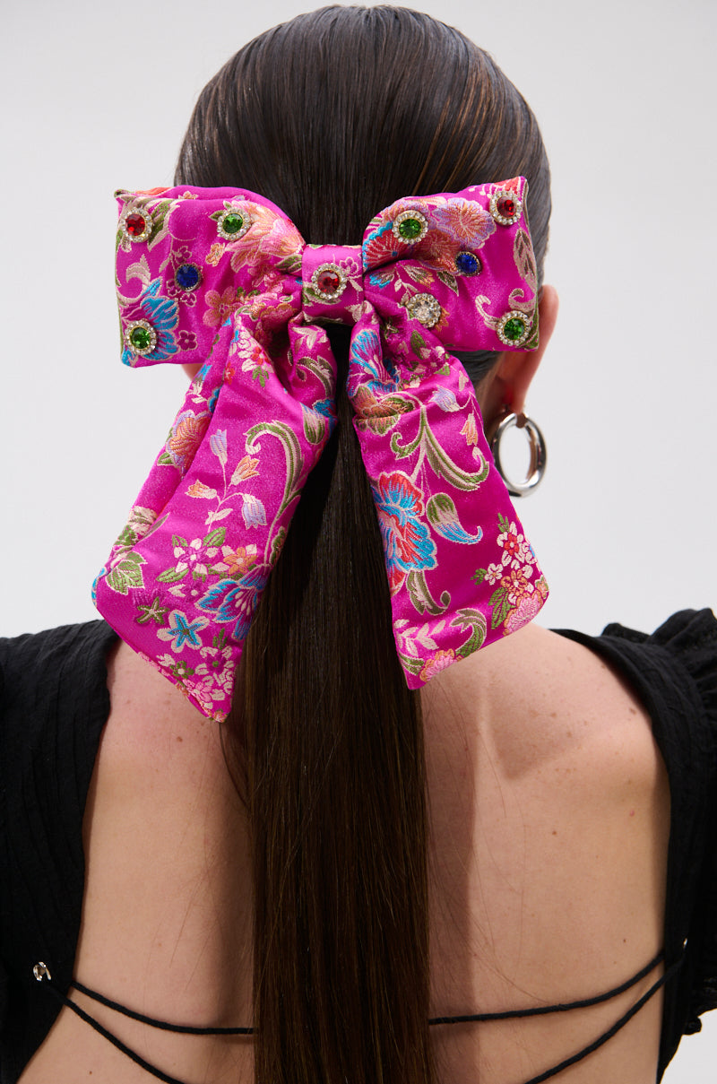 A person with long, straight brown hair wears the CICI DREAMS HAIR CLIP—a large, bright pink satin bow with floral patterns—tied at the back of the head. Dressed in black with hoop earrings, the standout bow adds charm to their look. 0