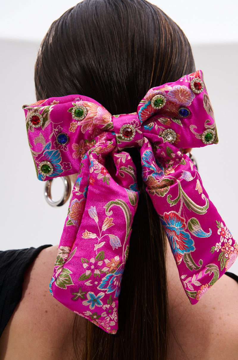 A person with long, straight brown hair wears the CICI DREAMS HAIR CLIP—a large, vibrant pink satin bow adorned with colorful floral patterns and sparkling gemstones—attached at the back of the head. 2
