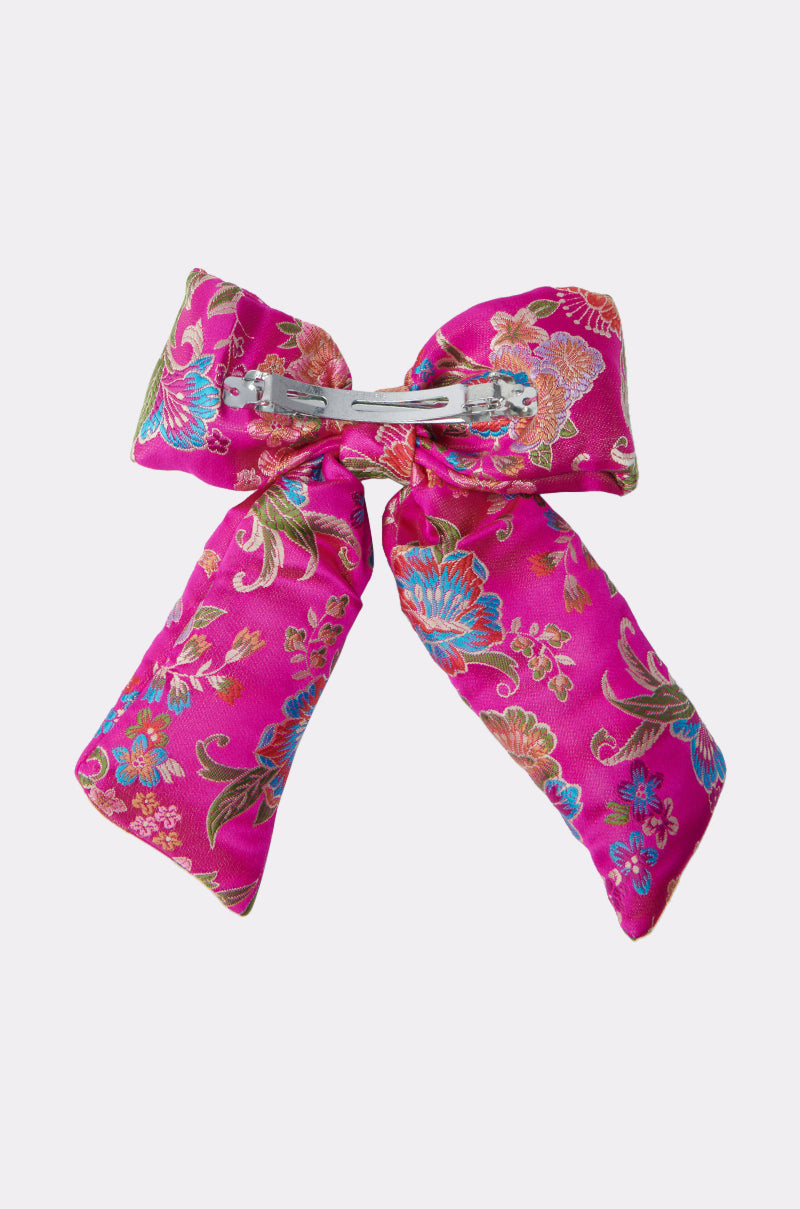 The CICI DREAMS HAIR CLIP is a vibrant pink satin bow with blue, green, and gold floral patterns, featuring a metal barrette backing. The silky bow is shown against a light background. 4