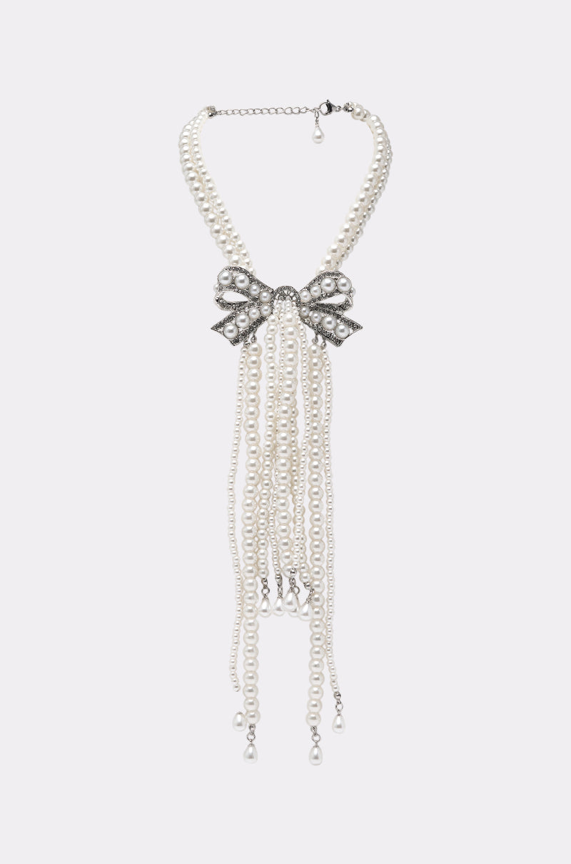 The SHANI PEARL NECKLACE features multiple pearl strands with a sparkling rhinestone bow centerpiece and elegant dangling pearl accents. 0