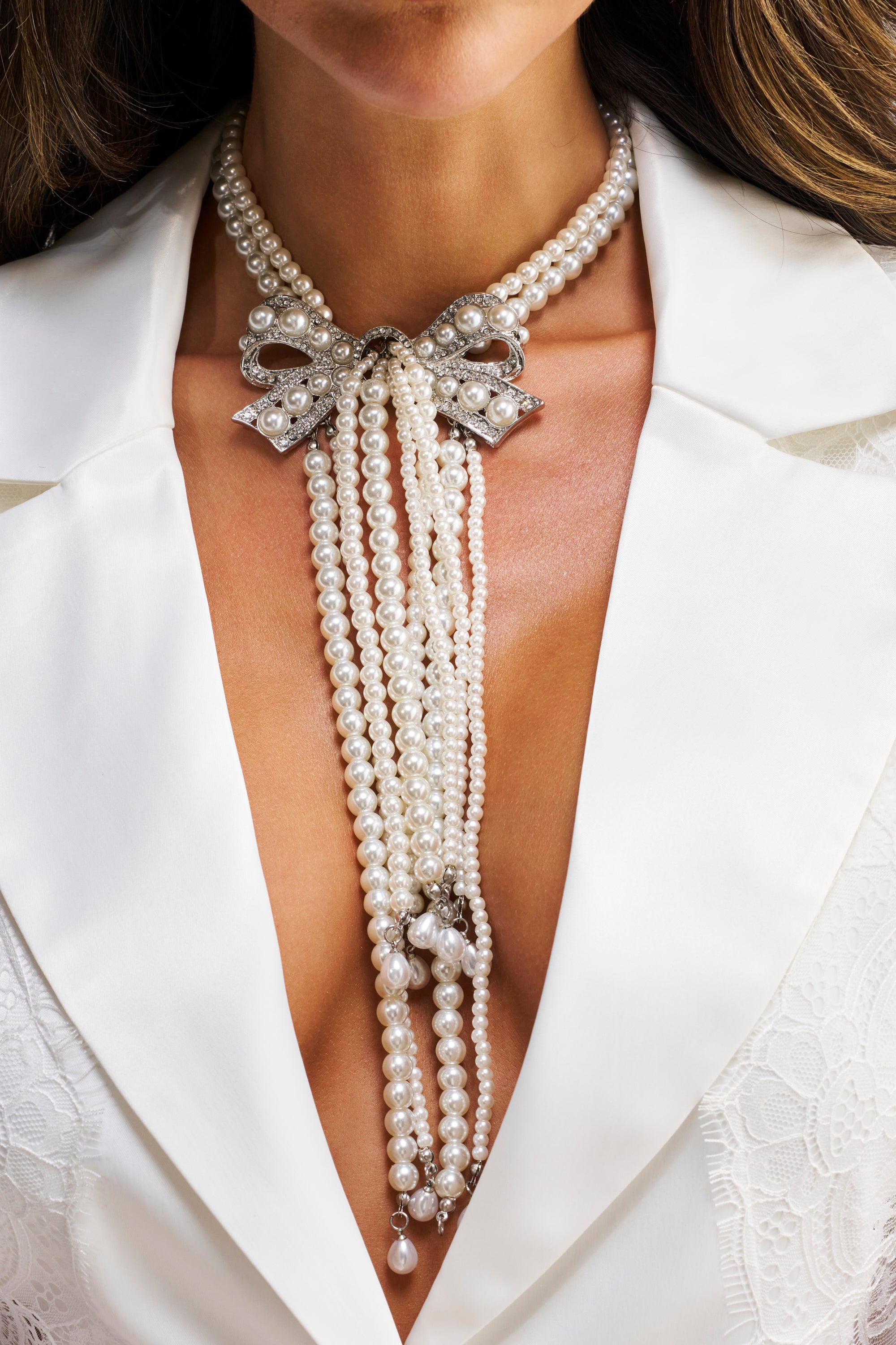 A woman wearing a white blazer and the SHANI PEARL NECKLACE, featuring a decorative bow clasp and cascading strands of pearls.