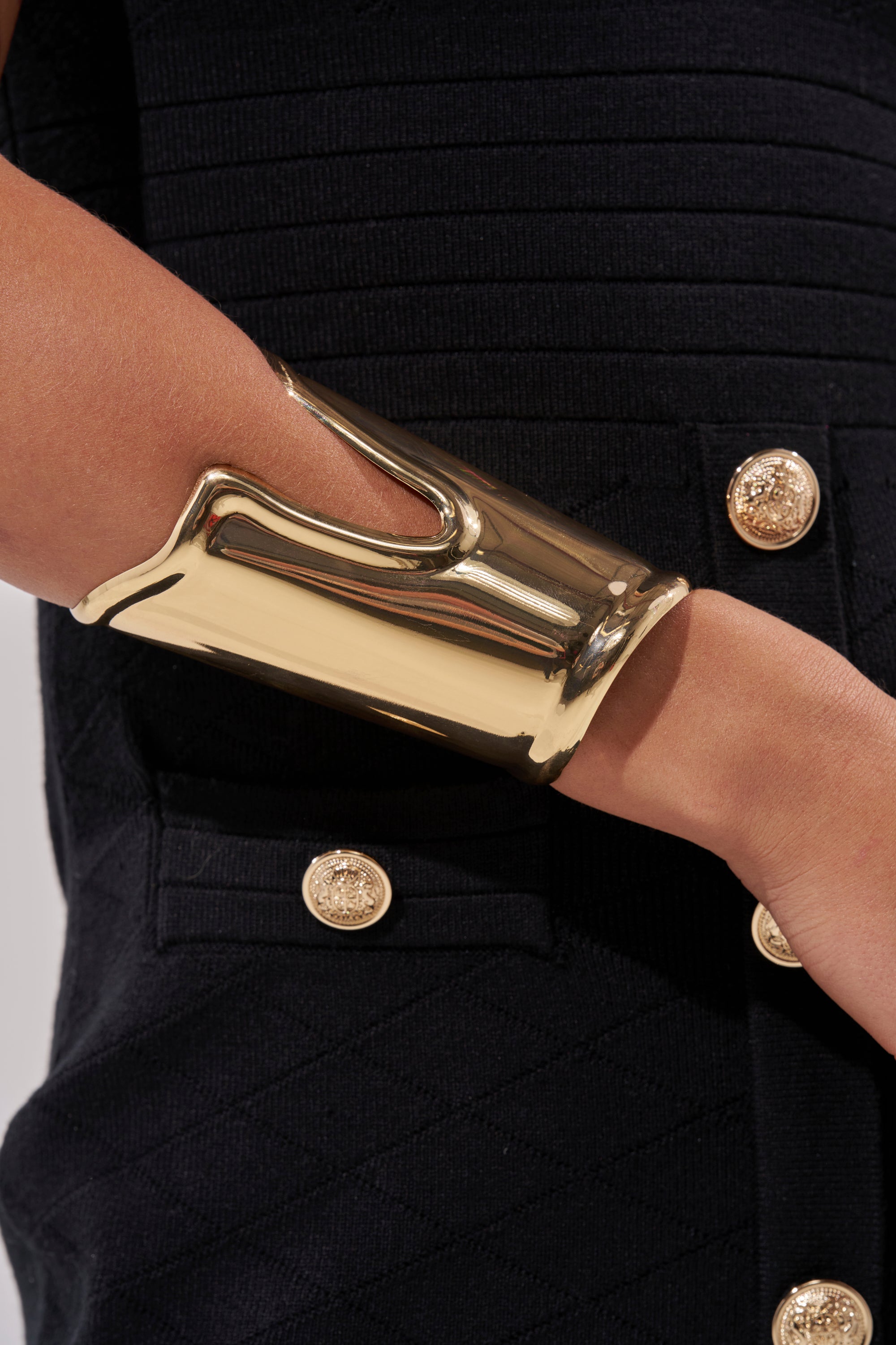 A close-up of a person wearing the OUTFIT COMPLETER CUFF, a statement gold bracelet, on their wrist, paired with a textured black garment detailed with gold decorative buttons. 1