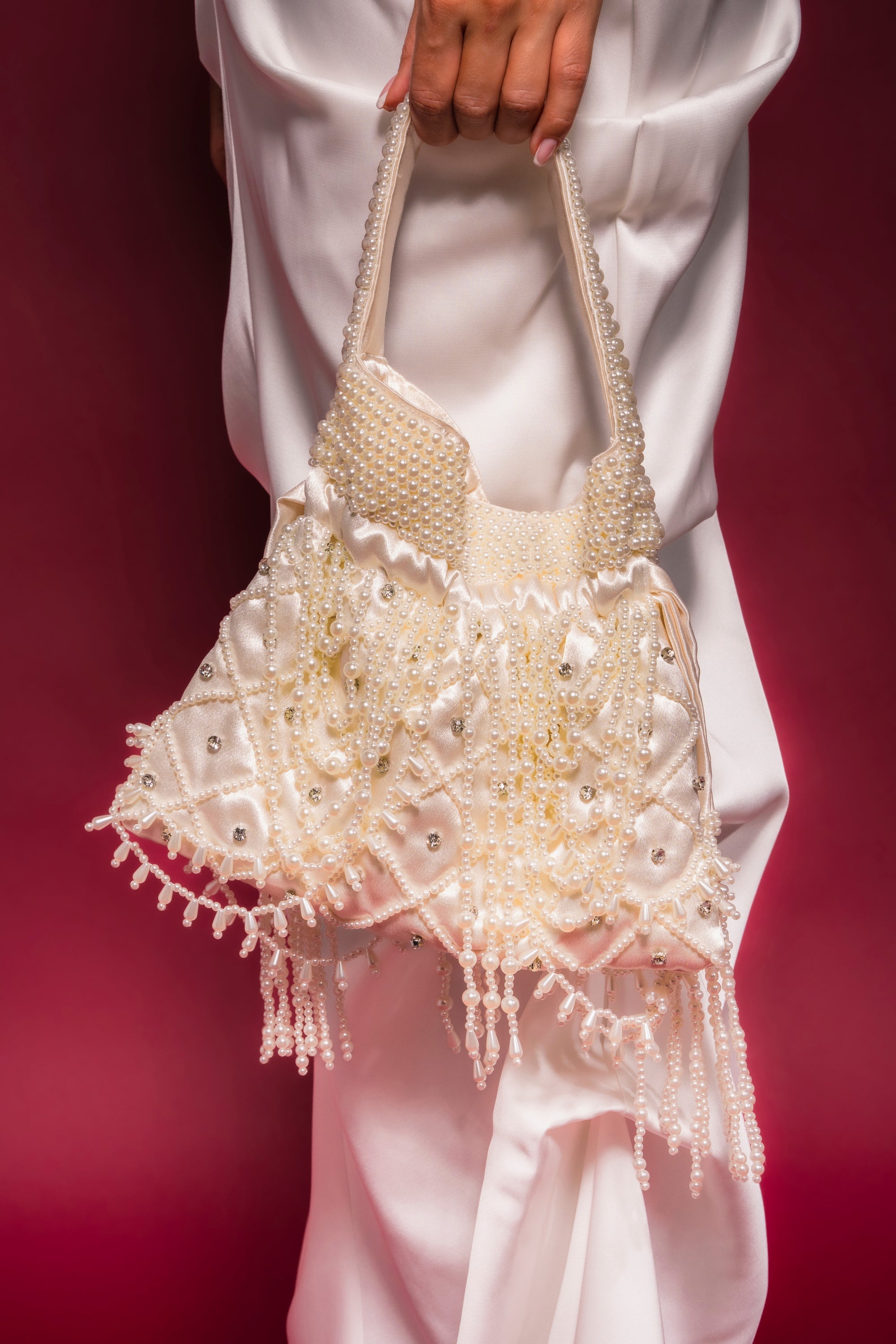 A person in white holds the POLLY PEARL PURSE, a cream-colored bridal bag with bead embellishments, fringe, and a textured handle, set against a reddish-pink background.