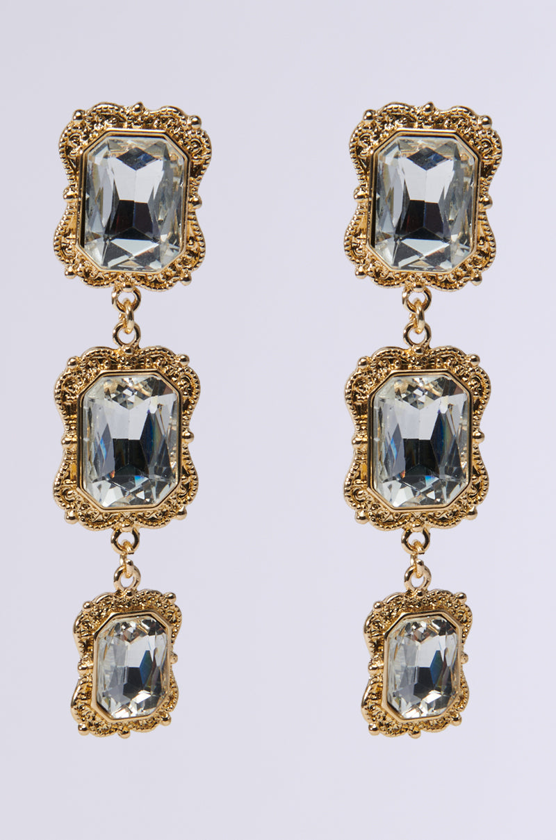 Discover the MIRROR MIRROR EARRING: an elegant pair of faux gold metallic drop earrings, each featuring three large rectangular faceted gemstones set in intricate gold designs, linked vertically. The minimalist white background accentuates their classic allure.