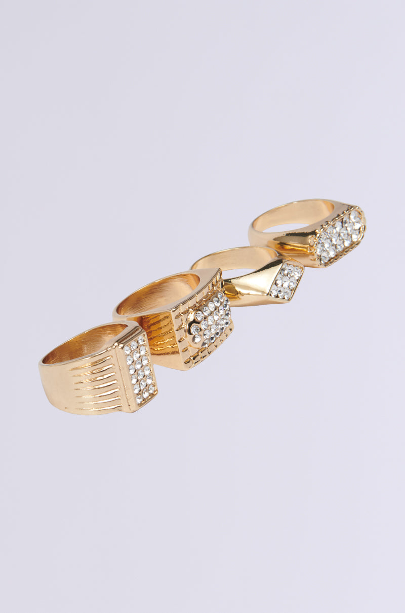 The EXPENSIVE TASTE RING SET includes four gold rings with ornate square and rectangular designs. Each ring serves as a geometric centerpiece, adorned with sparkling rhinestones against a plain background, exuding luxury and sophistication.