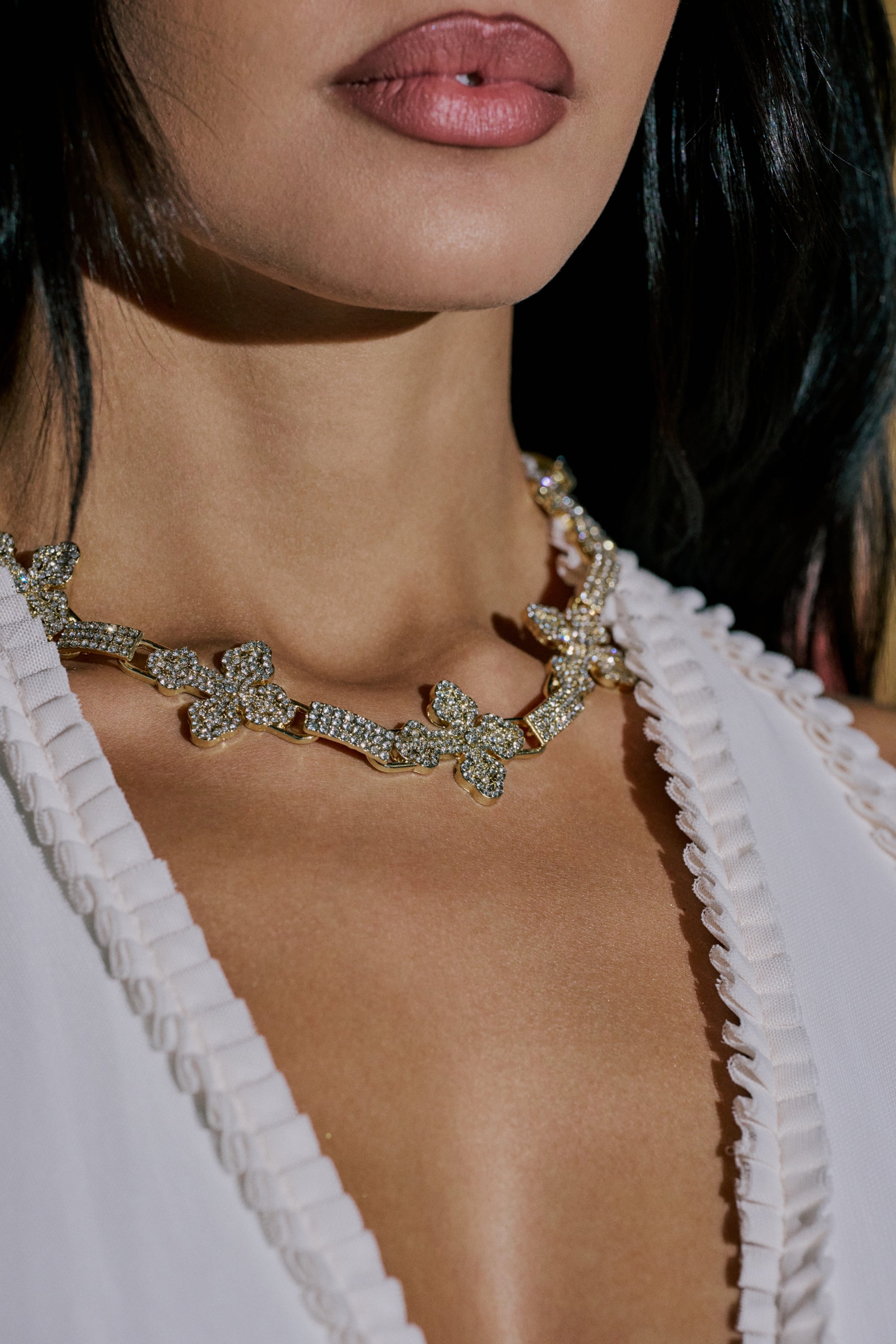 A close-up of a woman wearing the CROSS MY HEART NECKLACE, a gold chain adorned with sparkling cross pendants. She pairs it with a white v-neck top, her parted lips and dark hair elegantly framing her face. 1