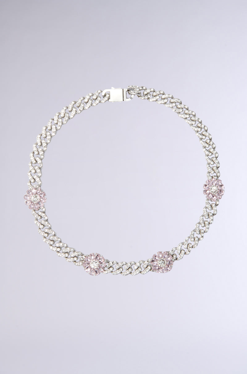 The PINK PETALS NECKLACE has a silver chain embellished with crystals and three pink floral-shaped charms with rhinestone centers, evenly spaced. Set on a gray background, its flower charms add elegance.