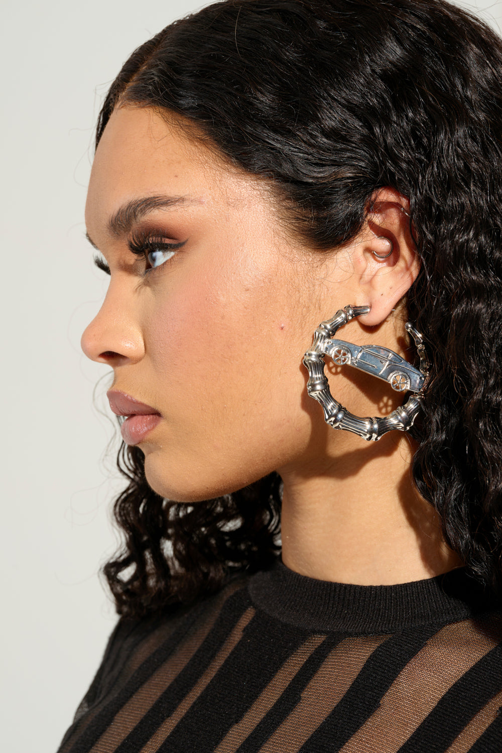 A woman with curly hair is wearing the RIDE OR DIE EARRING, which are oversized hoops crafted from a metallic material. She is dressed in a black top featuring sheer vertical stripes. Her makeup is natural, accentuating her eyes against the simple, light-colored background.