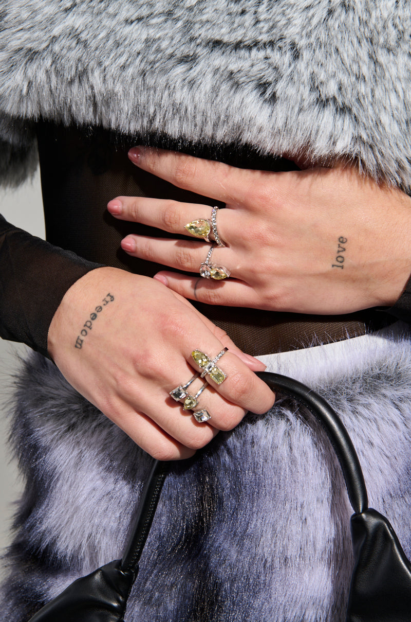 A close-up captures hands decorated with the STAY DRIPPY RING SET, showcasing opulent yellow gemstones and intricate spiral wrap-around designs in faux silver. The person is wearing a gray fur coat while holding a black strap, and tattoos spelling "peace" and "love" are visible on the fingers.