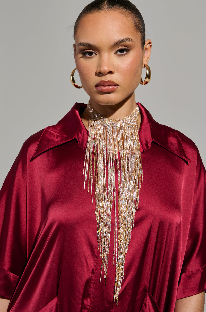 A person adorned with the IM A 10 NECKLACE—a shimmering, metallic faux gold fringe statement piece—paired with hoop earrings, dressed in a silky red outfit against a neutral background. The individual has a composed expression with their hair pulled back neatly. 1