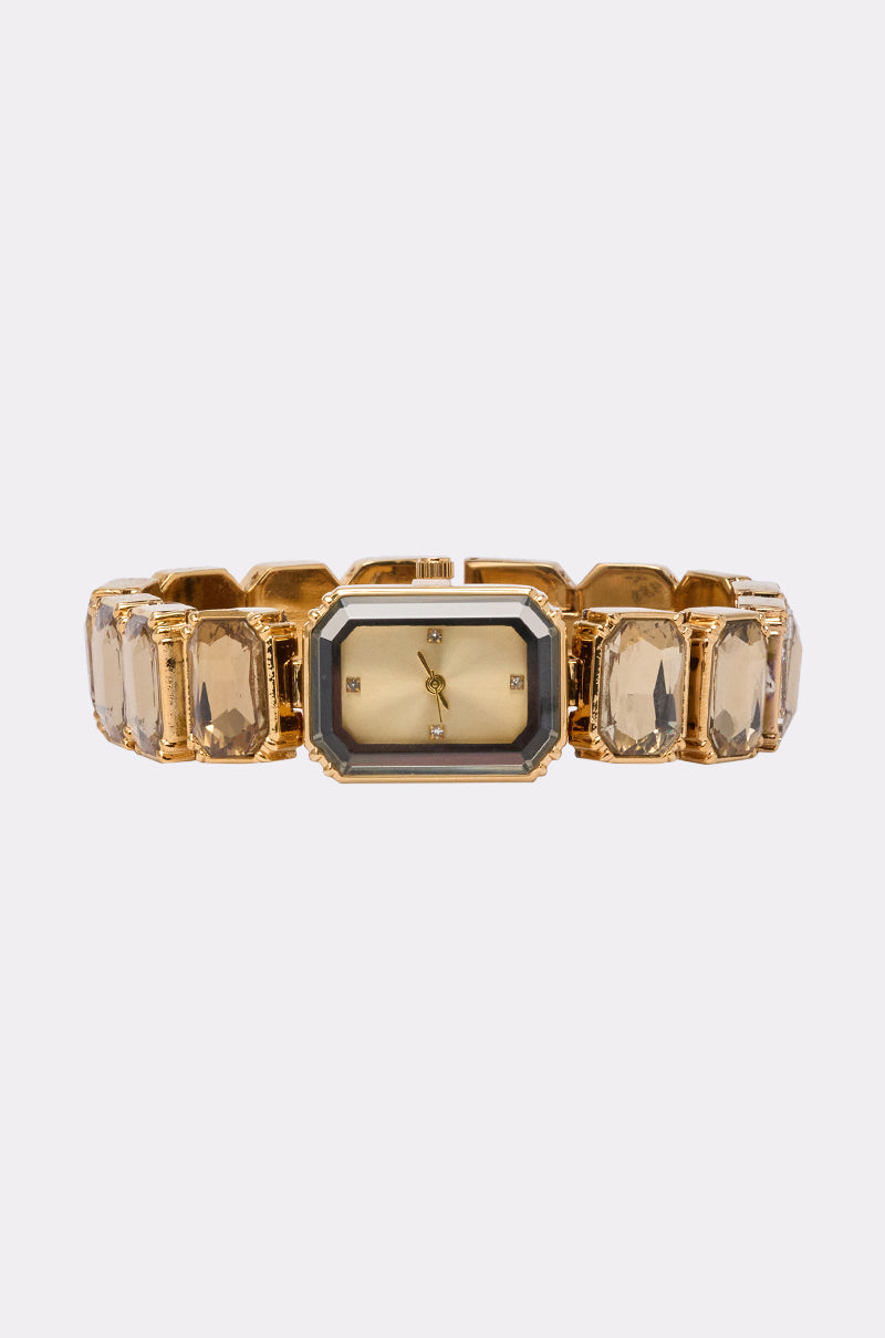 The SO GROOVY WATCH IN YELLOW features a gold-tone metal design with a rectangular face and clear gemstone accents on the band, shown against a plain white background. 1