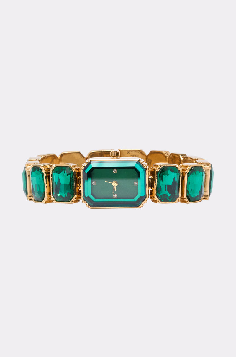 The SO GROOVY WATCH IN GREEN features a gold wristband, a rectangular emerald green face, rhinestone hour markers, and green gemstone accents on the band, displayed on a plain white background. 0
