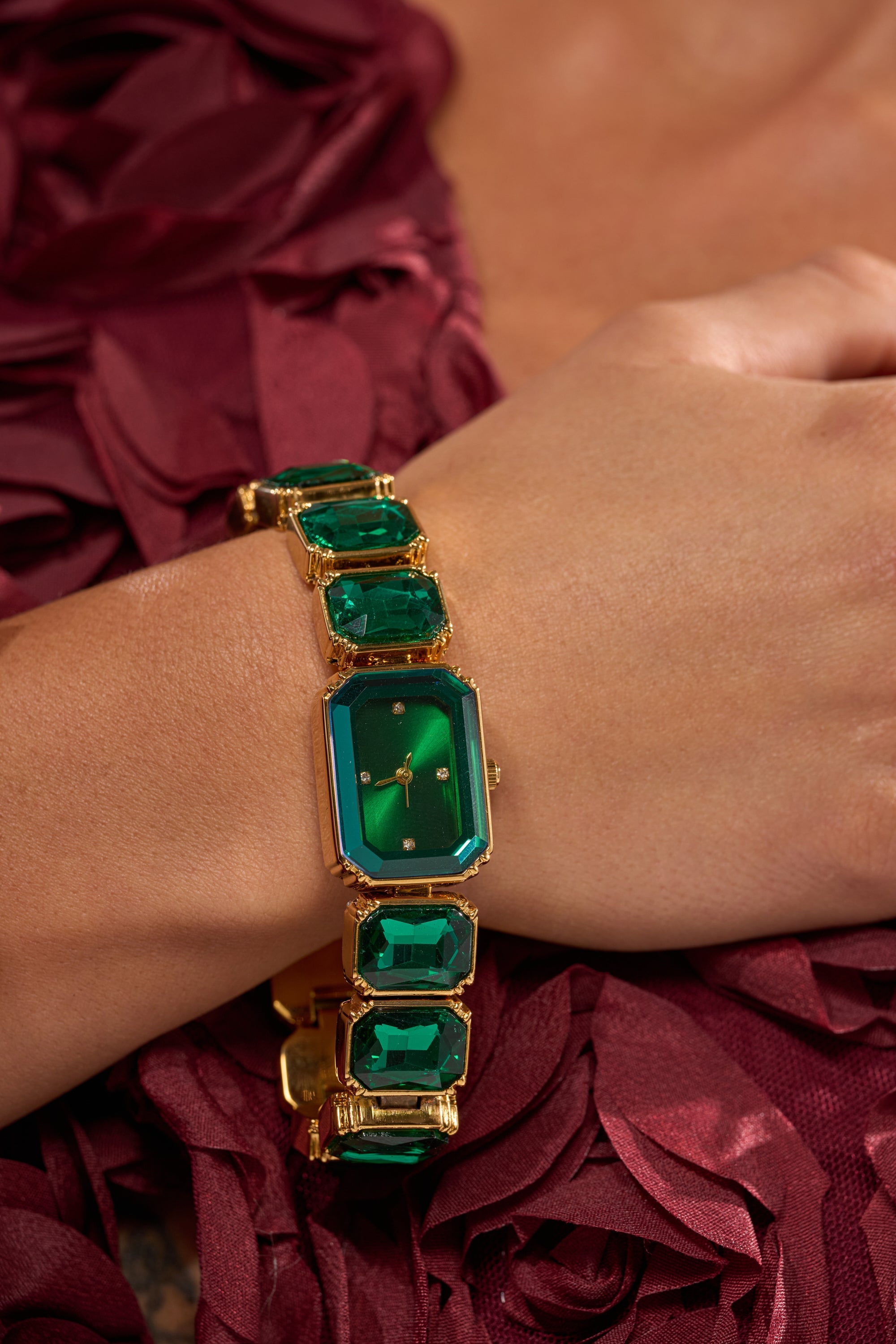 A hand wearing the SO GROOVY WATCH IN GREEN with gem links rests on burgundy fabric featuring textured floral patterns. 1