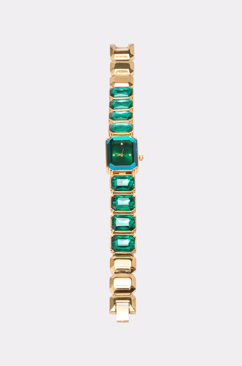 The SO GROOVY WATCH IN GREEN by AKIRA Label features a gold-tone design with a square green face and a band decorated with rectangular green gemstones, shown against a plain light gray background. 2