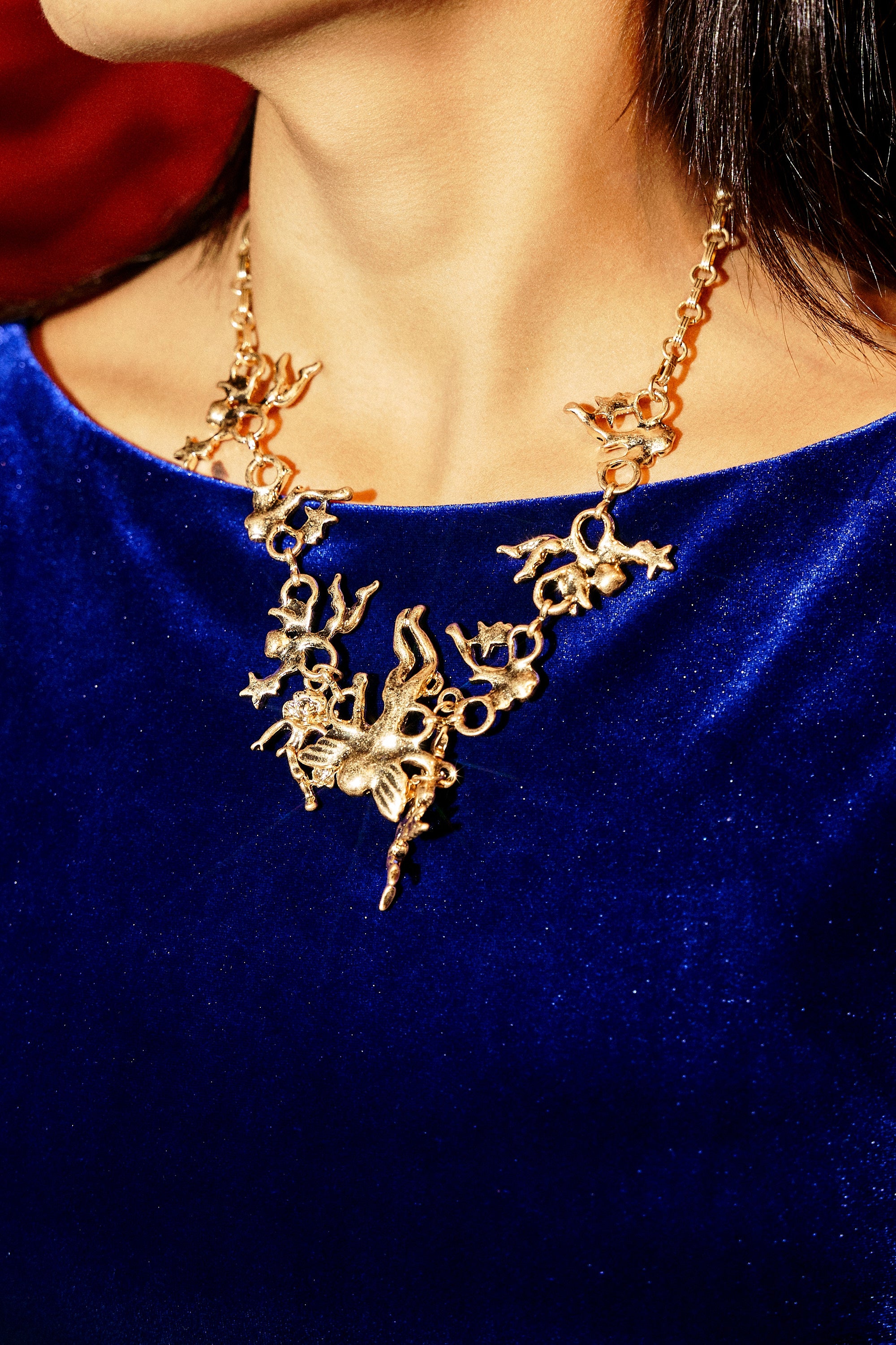 A person in a royal blue velvet top wears the ANGELIC NECKLACE, featuring gold-tone cherub charms and vintage-inspired details, photographed from the shoulders up.