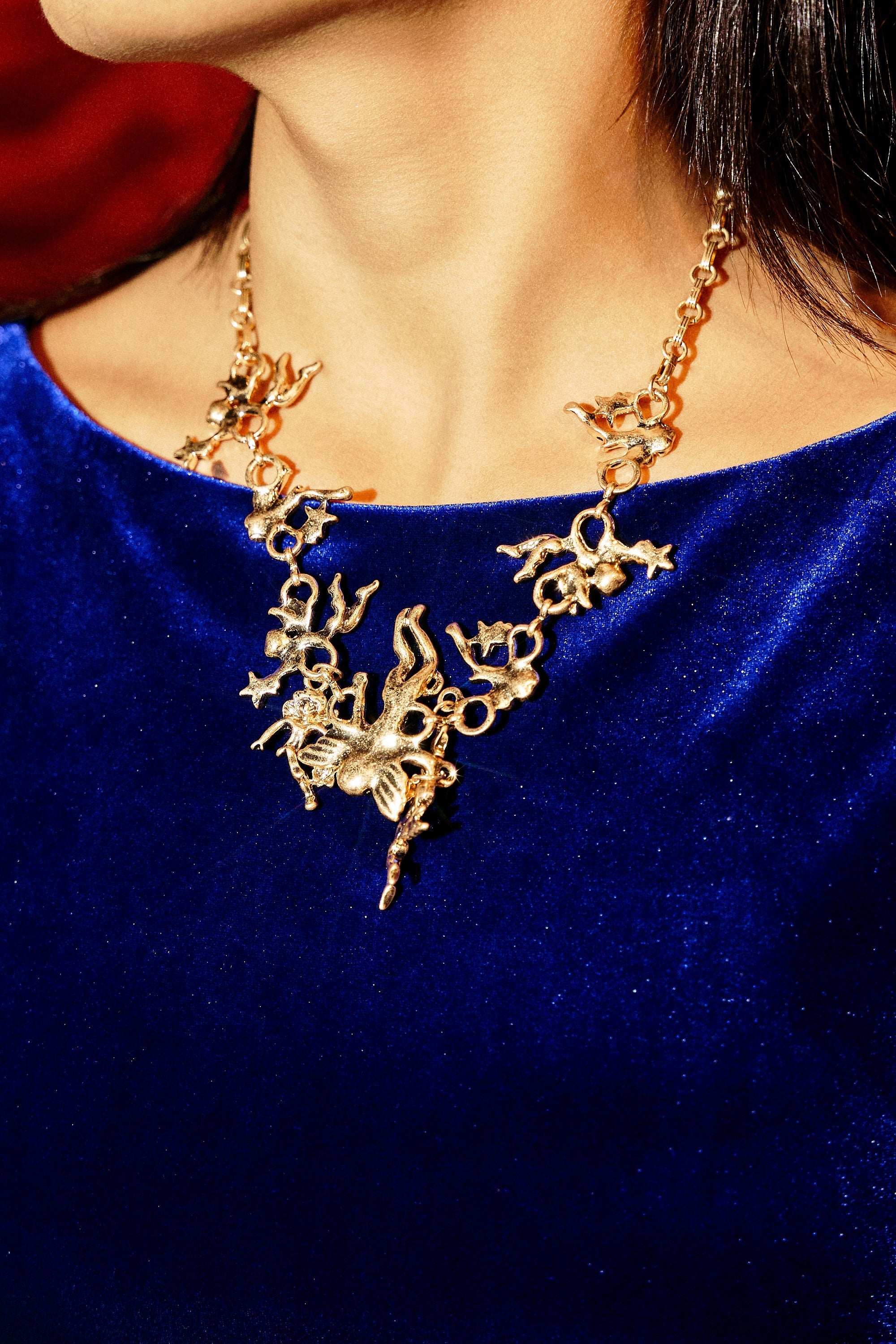 A person in a royal blue velvet top wears the ANGELIC NECKLACE, featuring gold-tone cherub charms and vintage-inspired details, photographed from the shoulders up. 1