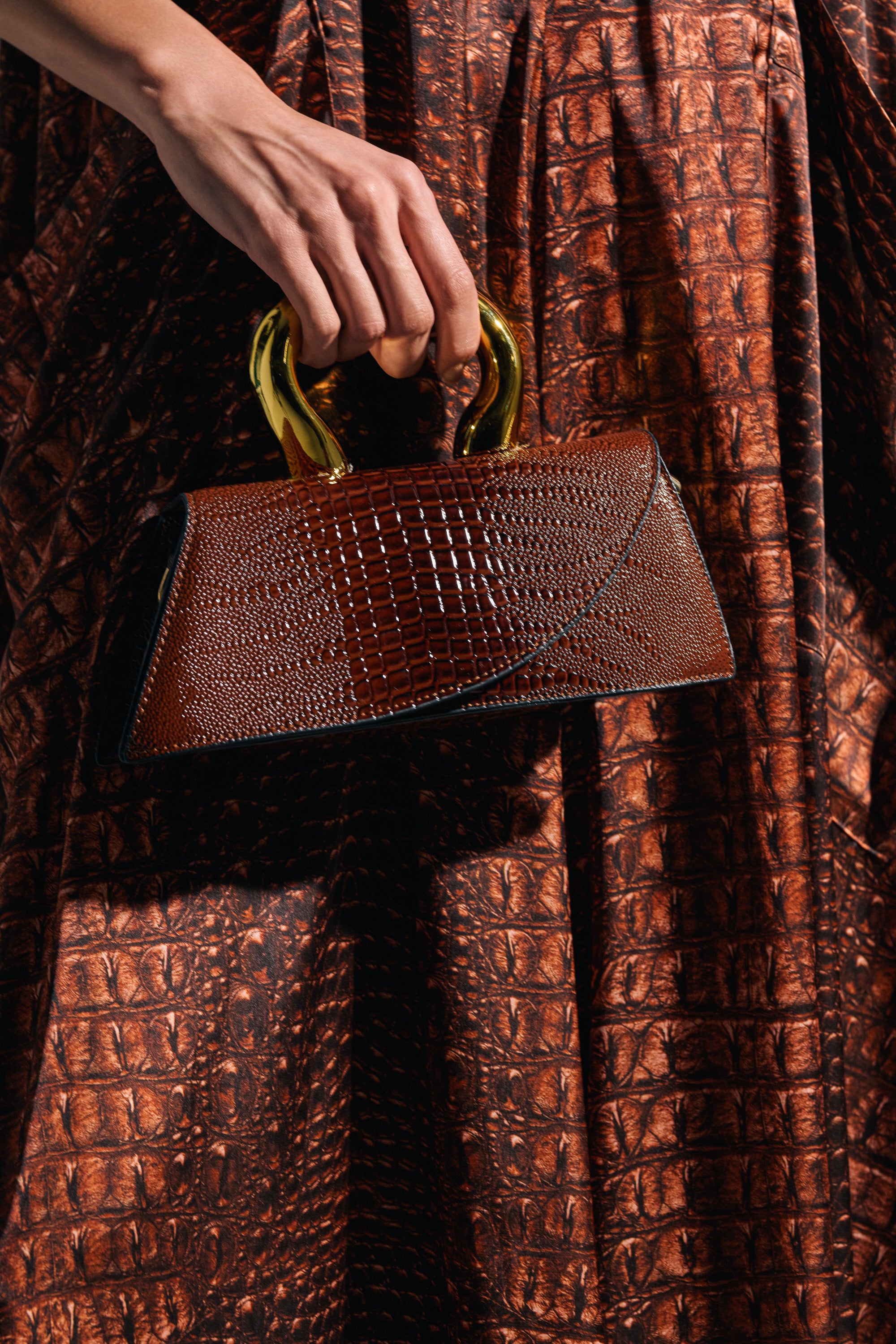 A person holds the DOESNT DESERVE ME PURSE, a small brown embossed reptile bag with gold handles, matching their crocodile-patterned brown skirt or dress.