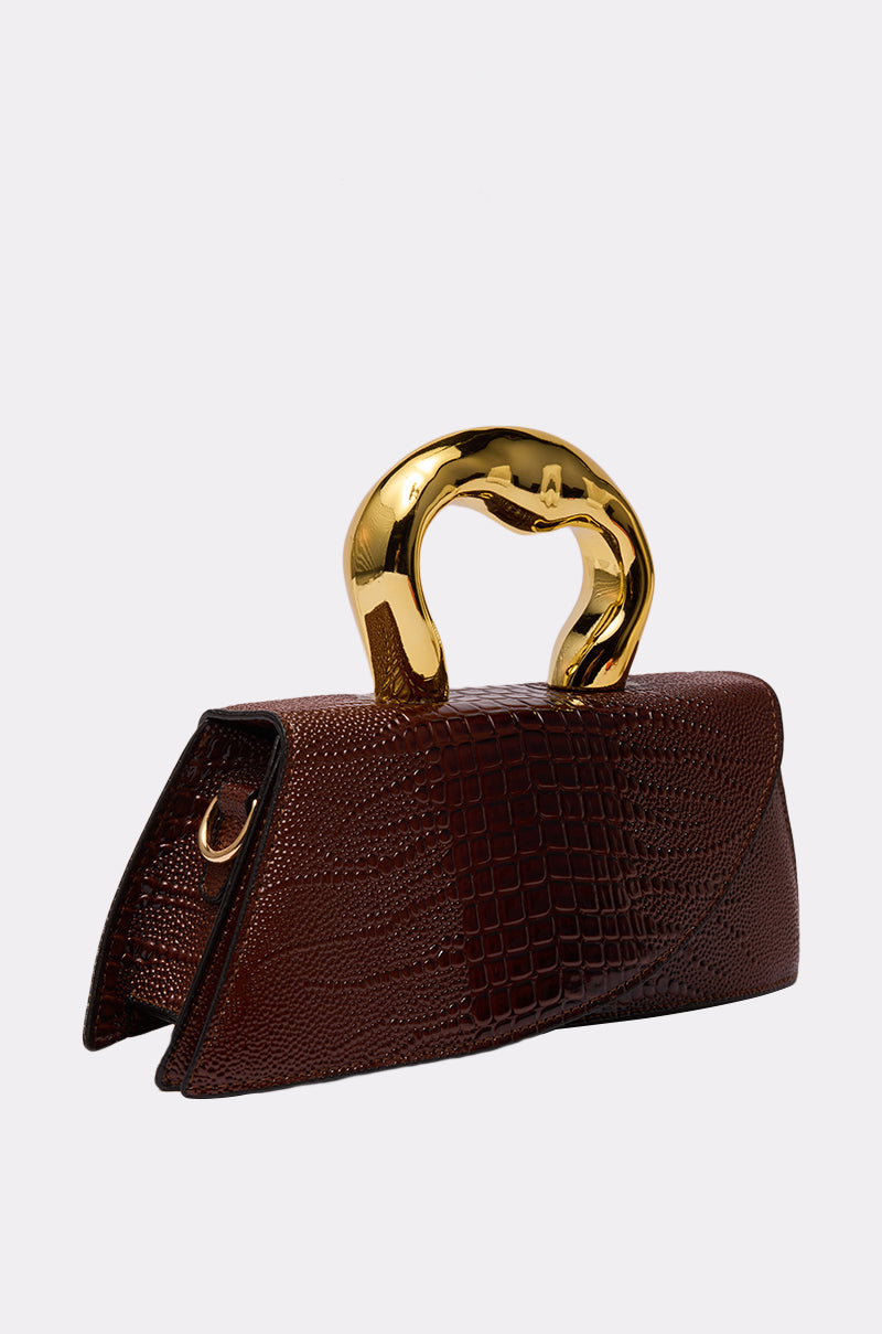 The DOESNT DESERVE ME PURSE is a brown structured crossbody bag with a reptile pattern and a shiny, curved gold handle, shown against a plain white background.