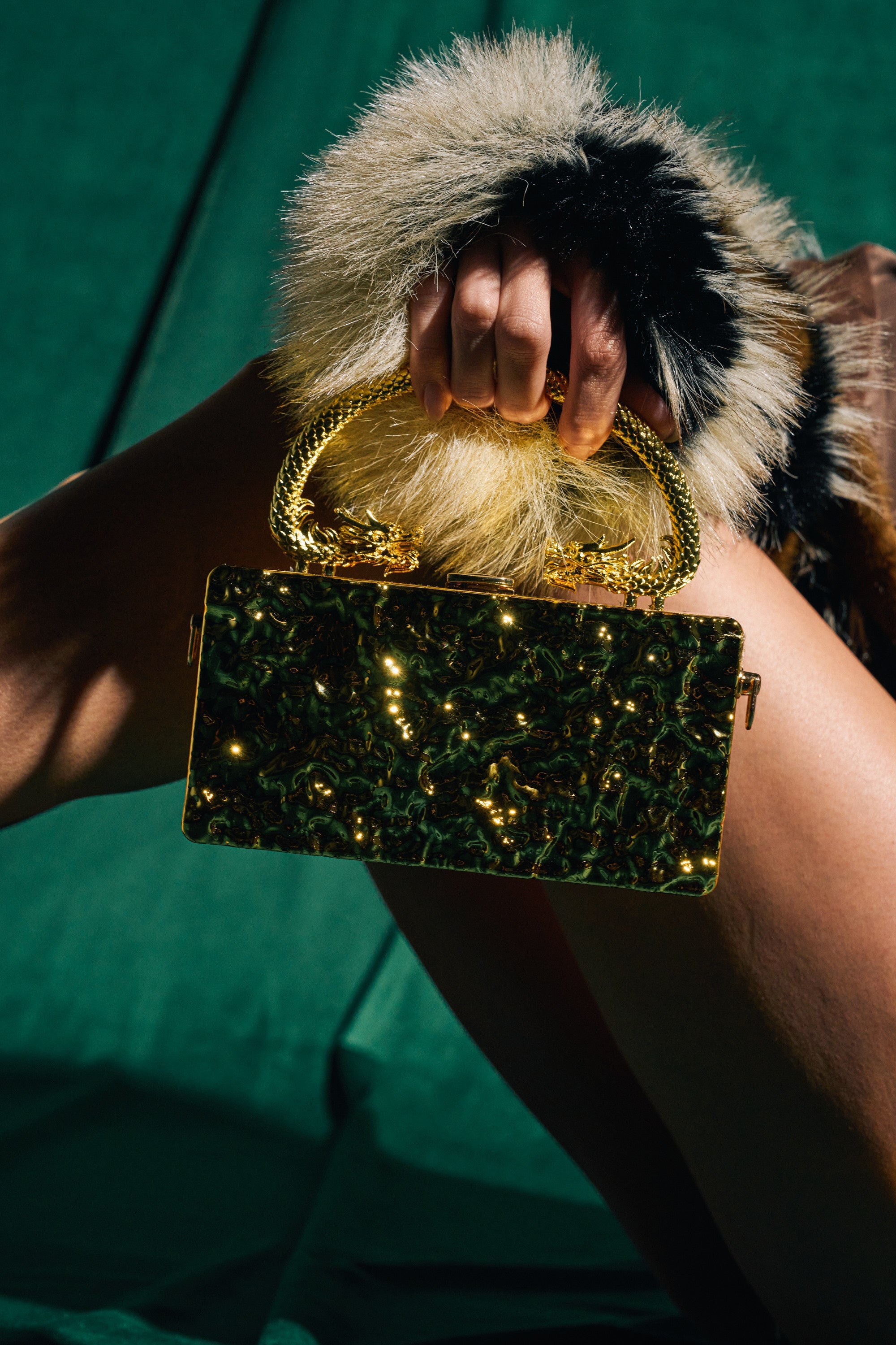 A person holds the MY KINGDOM CHROME PURSE, featuring a metallic gold finish and ornate dragon handle, while wearing a garment with fluffy black and beige fur cuffs against a green fabric background. 0