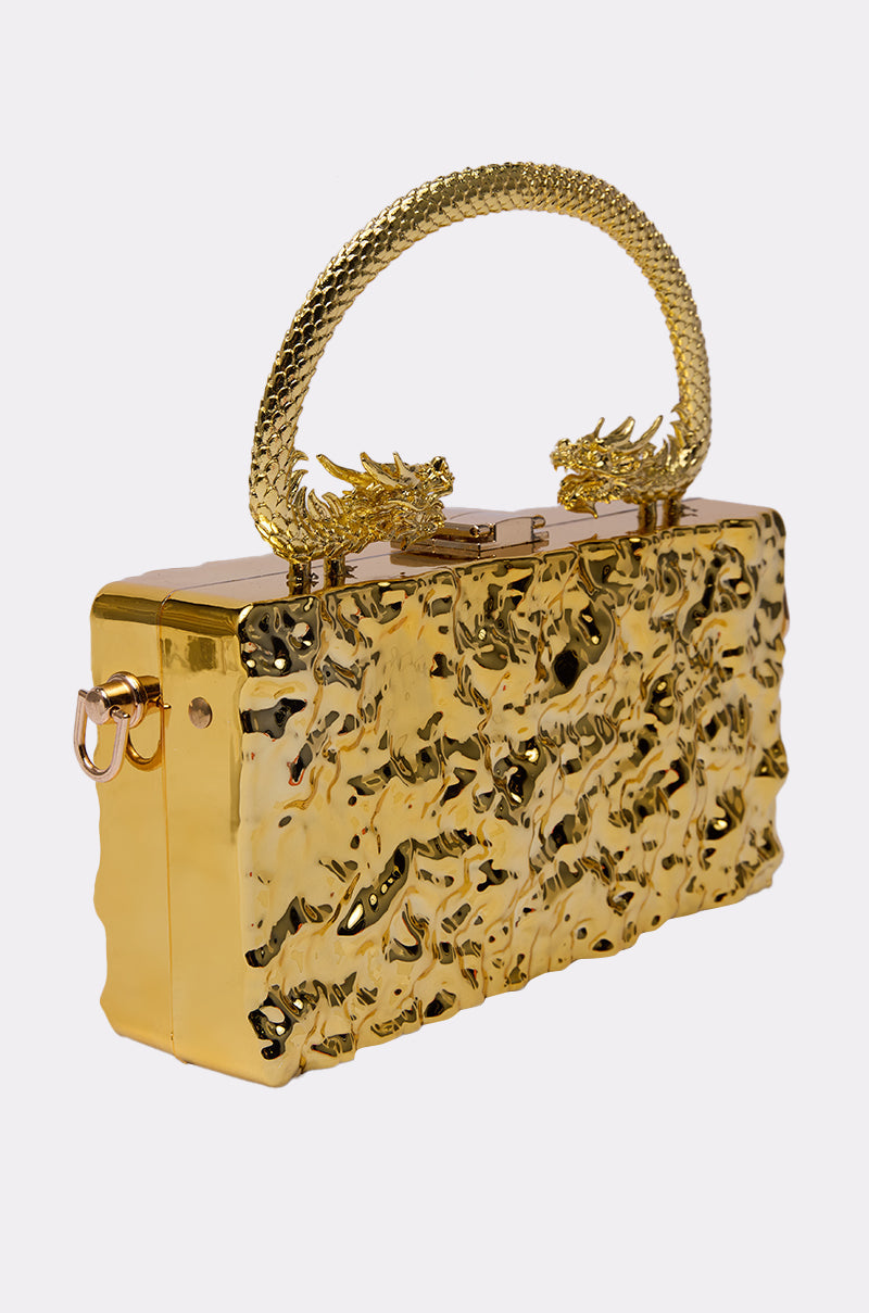 The MY KINGDOM CHROME PURSE is a rectangular, metallic gold bag with a shiny finish, dragon handle, and dragon head clasps, displayed on a plain white background. 2