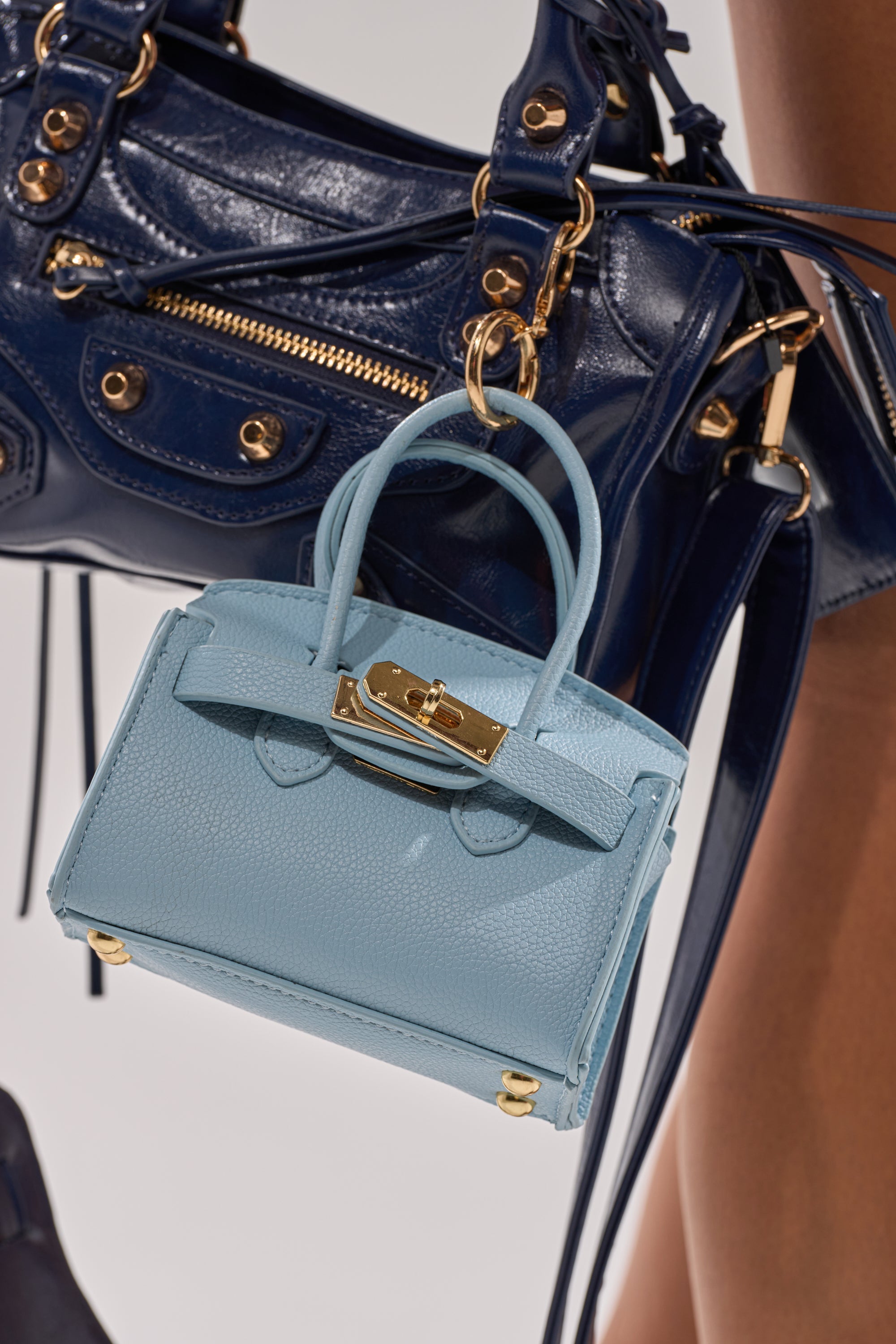 A close-up of a navy blue leather handbag with gold hardware, featuring the NEVER TOO BIG BAG KEYCHAIN—a small, light blue purse-shaped accessory with gold details—attached to it. A person’s leg appears in the background. 0