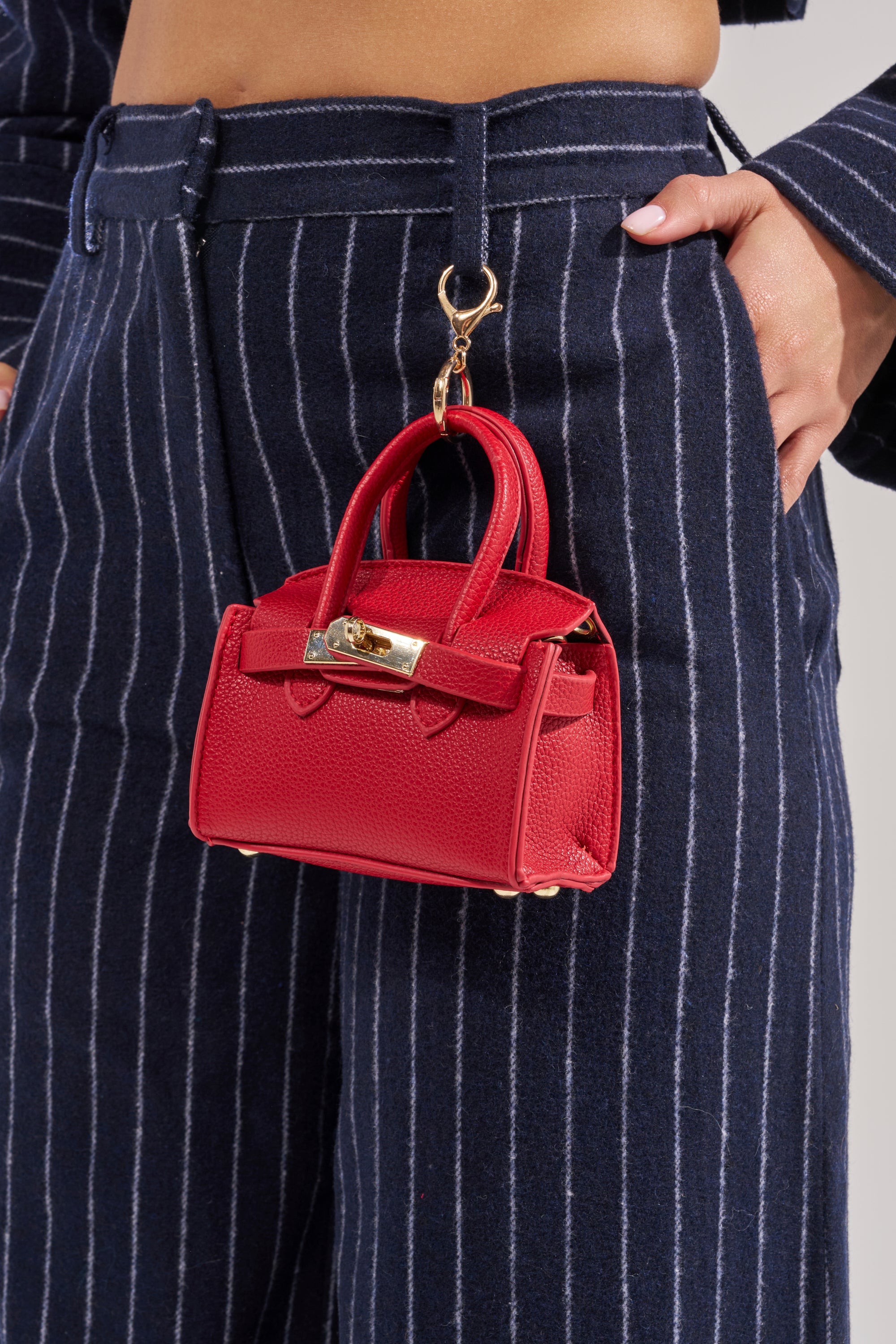 A person wearing navy blue pinstriped pants holds the NEVER TOO BIG BAG KEYCHAIN with gold hardware, attached to their belt loop by a clasp. 0
