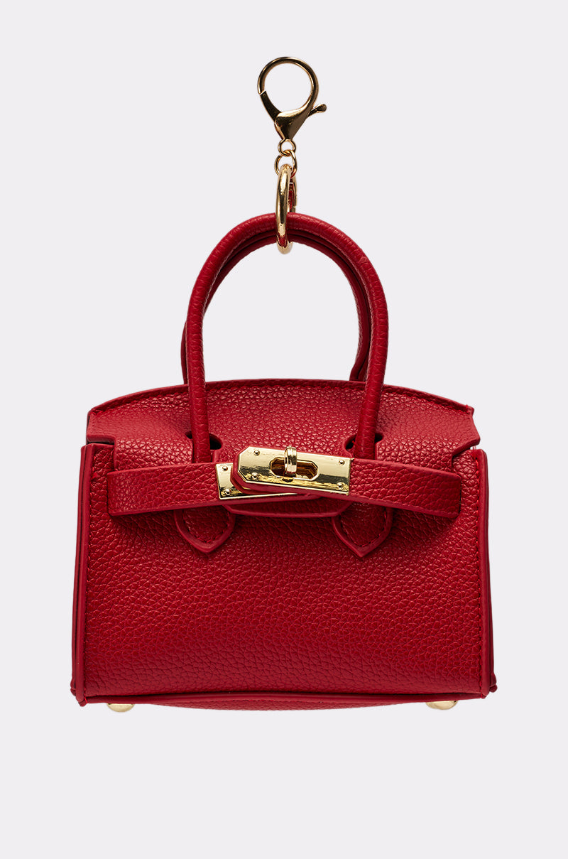 The NEVER TOO BIG BAG KEYCHAIN is a small red faux leather mini purse with gold hardware, two handles, a clasp closure, and a gold keychain clip. Stylish and perfect as an accessory; photographed on a plain light background. 1