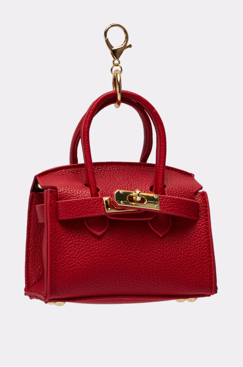 The NEVER TOO BIG BAG KEYCHAIN is a small red faux leather purse keychain with gold hardware, a textured surface, two handles, and a clasp, shown isolated on a light grey background. 2