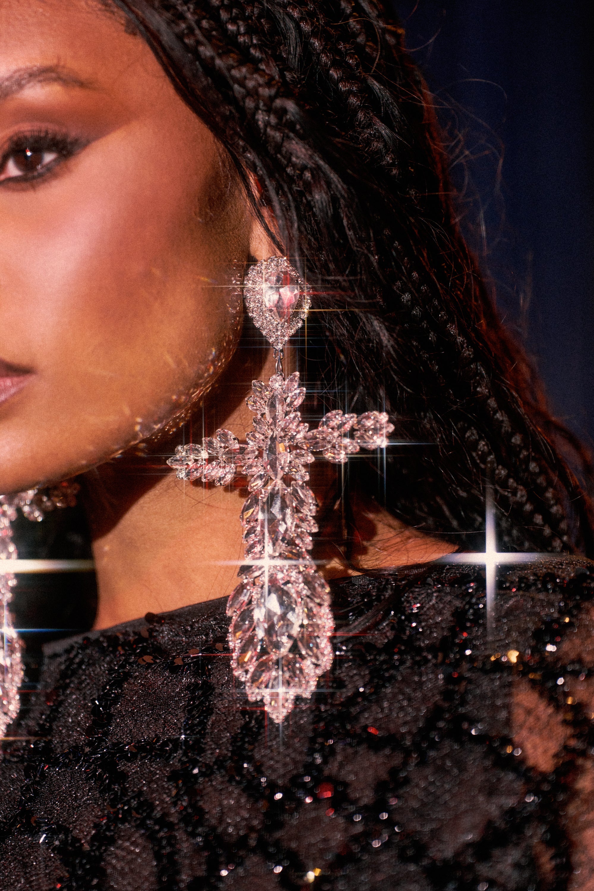Close-up of a woman wearing the WITH GRACE EARRING—oversized, crystal-adorned crosses—paired with a sequined black top. Her braided hair and the earring's sparkle under lights create a dazzling, glamorous vibe. 0