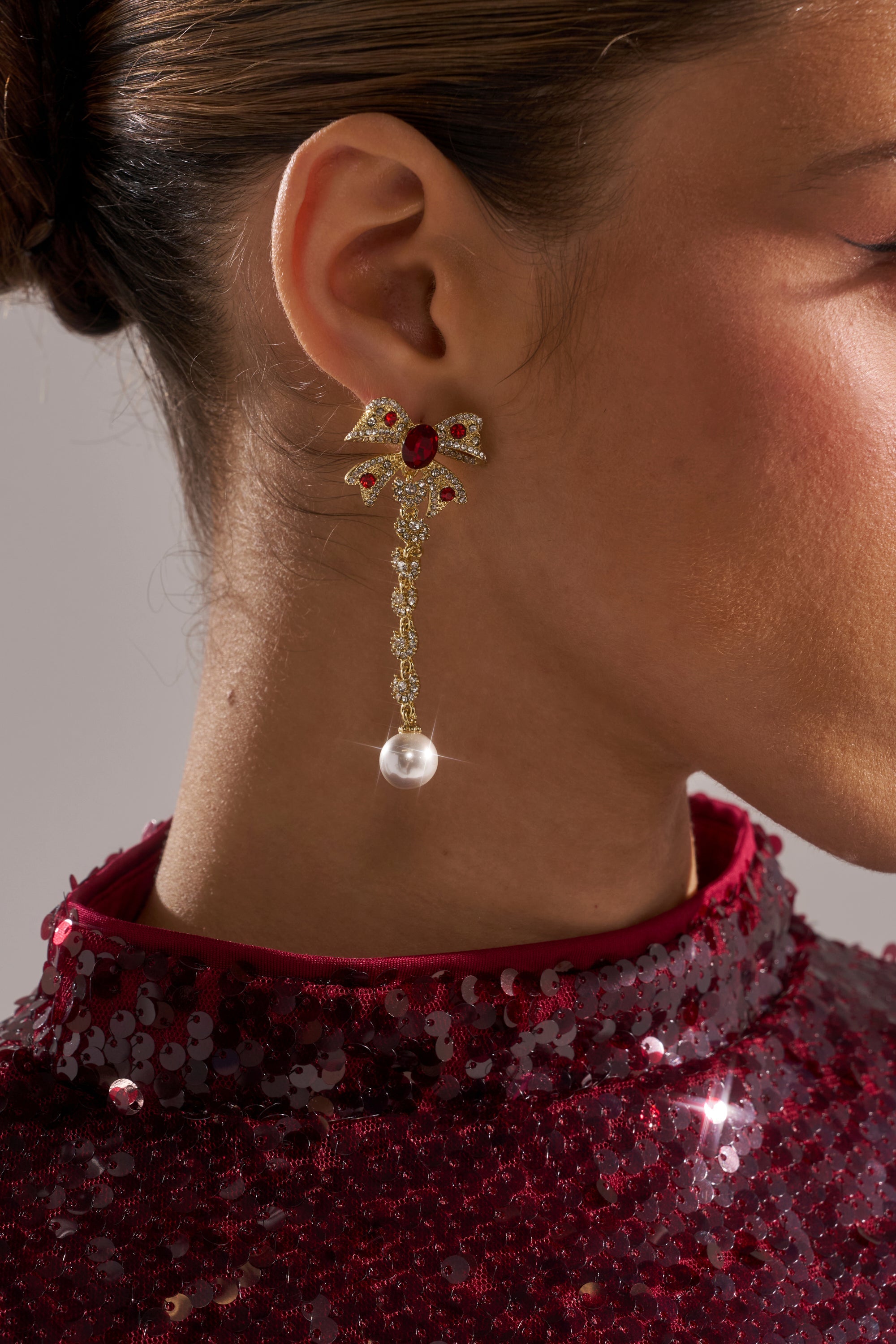 Close-up of a woman wearing TIE WITH A BOW EARRING featuring red gemstones, a dangling chain, and a faux pearl drop. She is dressed in a sequined burgundy garment. 1
