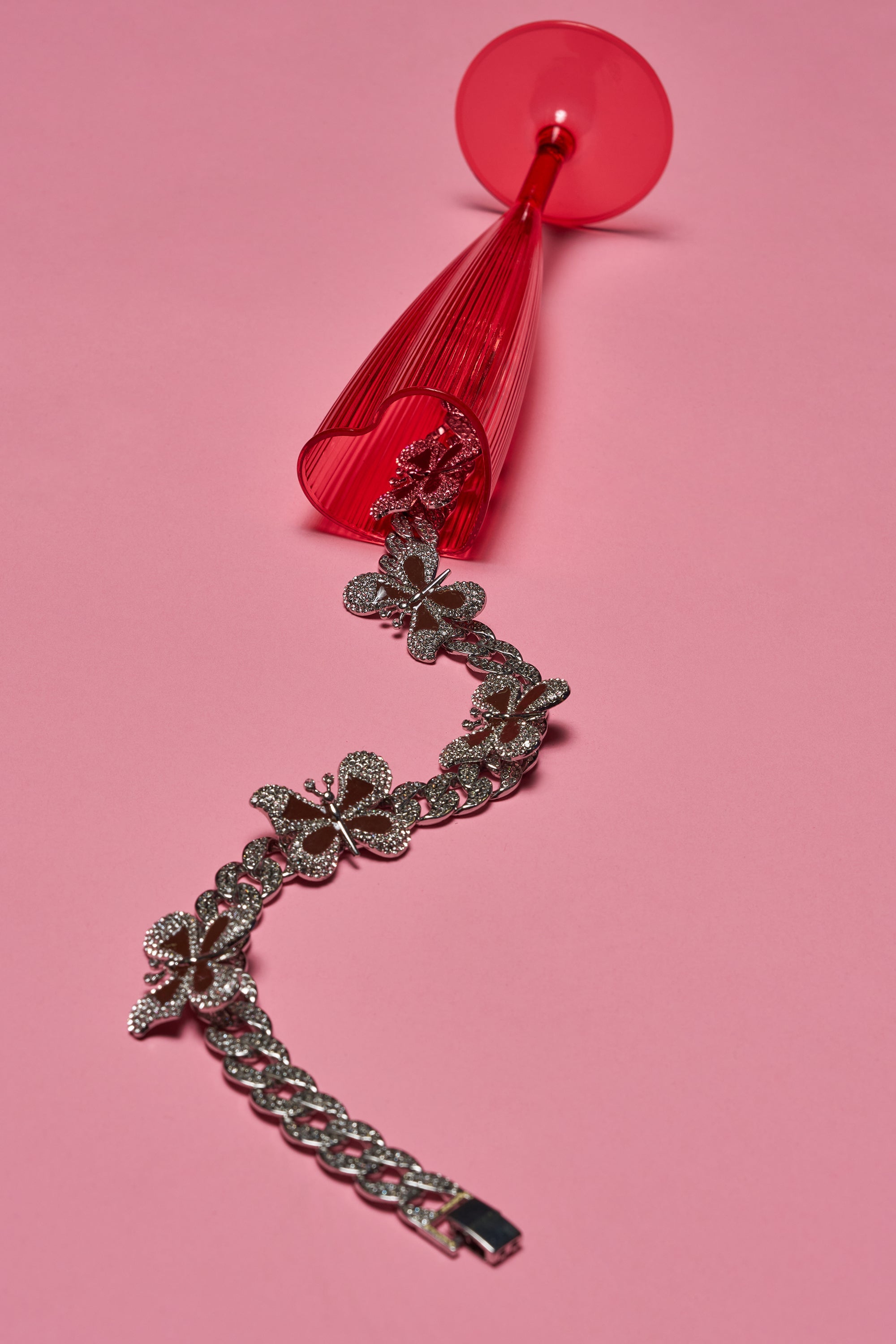 The LUXE BUTTERFLY NECKLACE with red enamel butterfly charms spills out of an overturned red wine glass on a pink background. 0