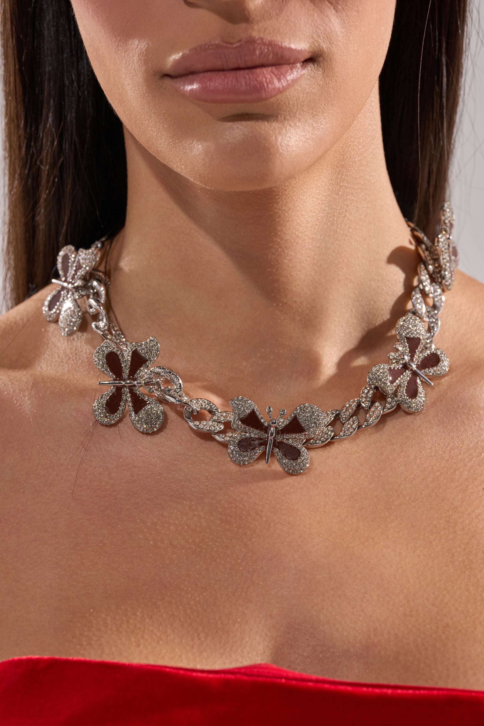 Close-up of a woman in a red strapless top wearing the LUXE BUTTERFLY NECKLACE, a chunky rhinestone Cuban link chain with sparkling butterfly and flower charms. She has straight brown hair. 2