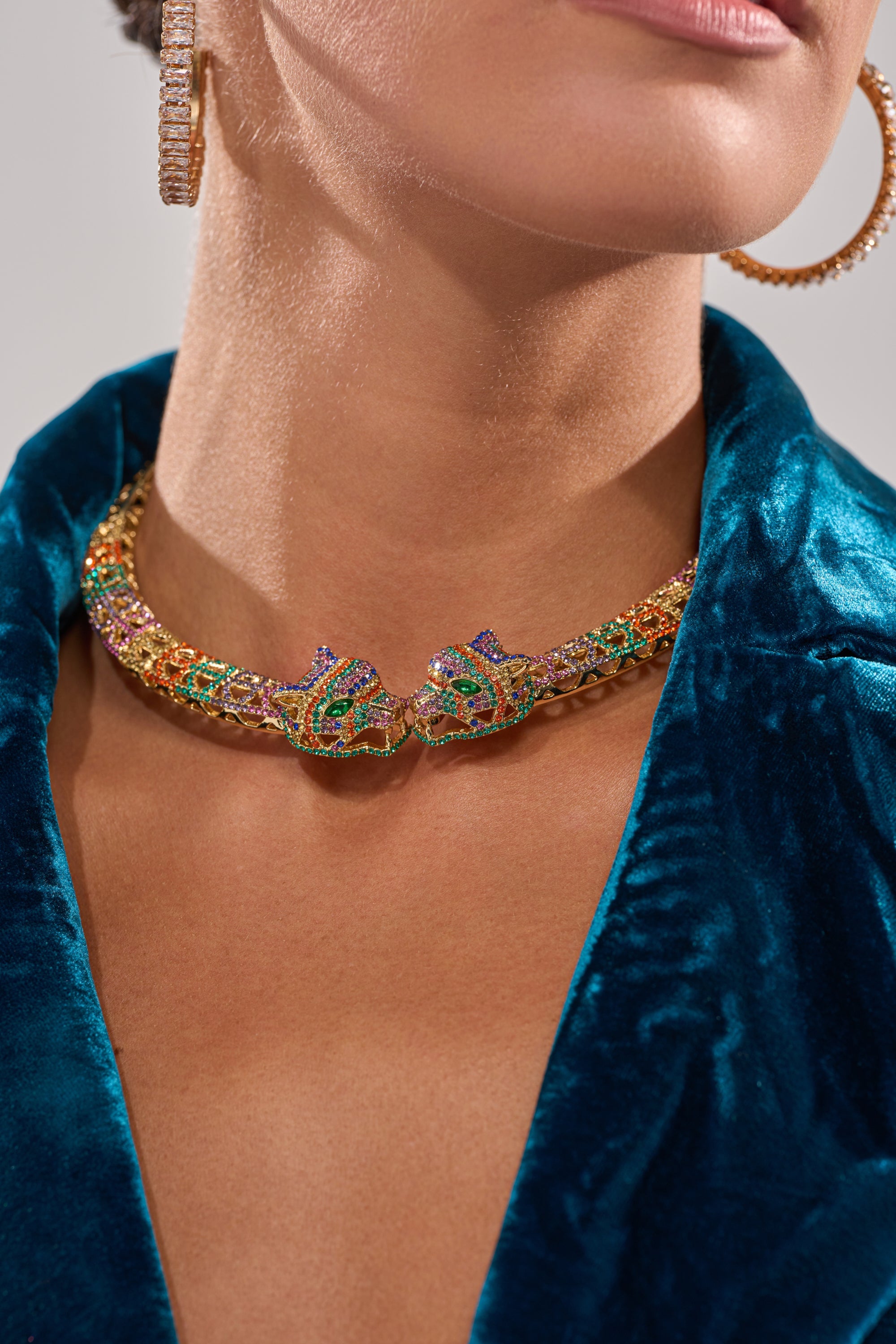 Wearing a blue velvet garment, the person showcases the LUXE JAG NECKLACE, featuring two ornate jaguar heads facing each other, paired with a large hoop earring. Only the lower face and neck are visible. 2
