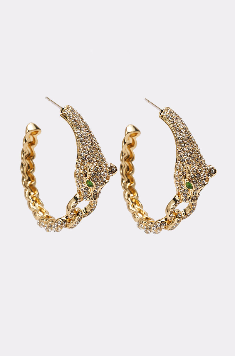 The LUXE JAG EARRING is a pair of gold hoop earrings shaped like leopards, featuring a jaguar head design with sparkling rhinestones and green gem eyes, shown on a plain white background. 1