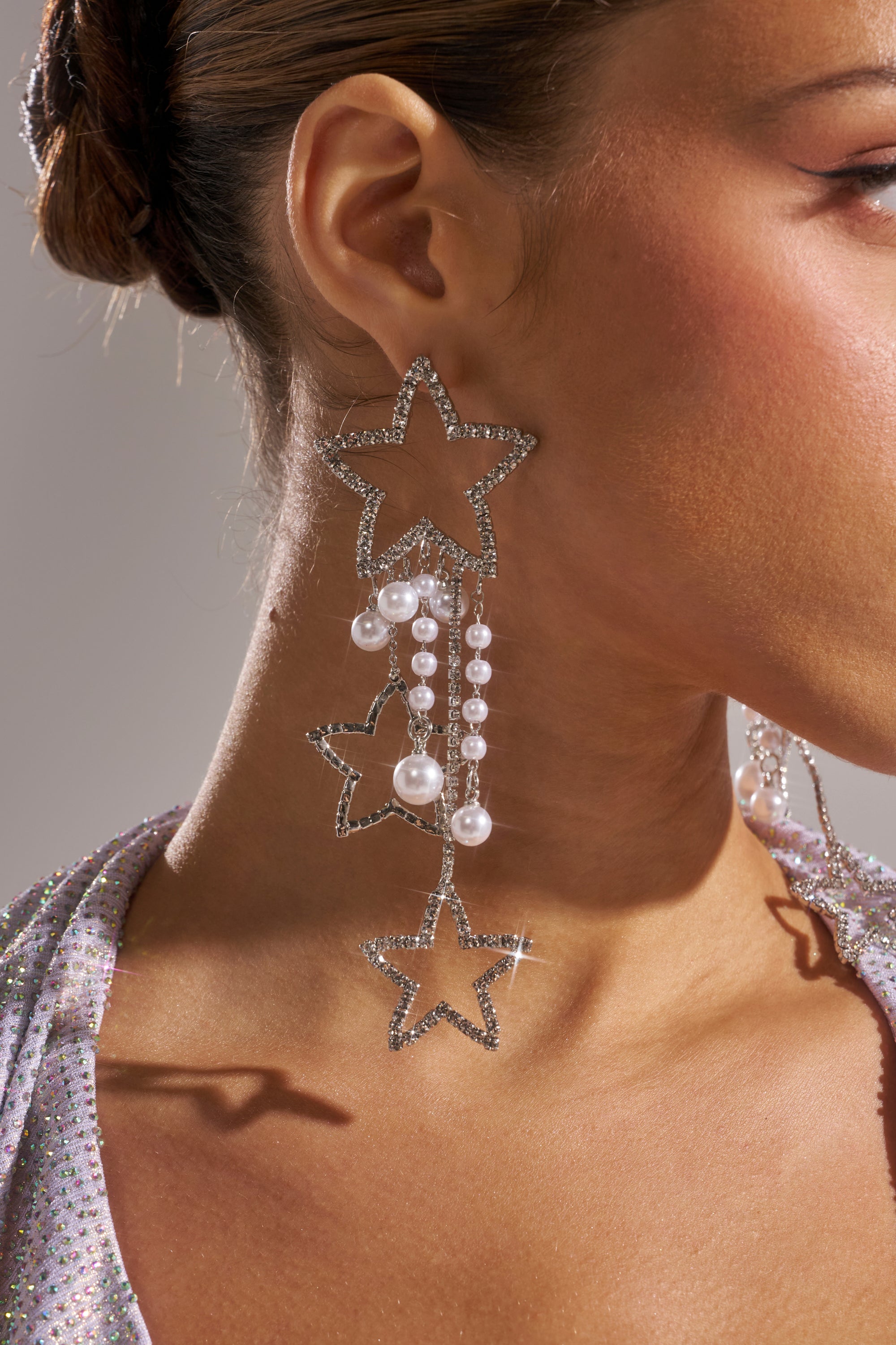 A close-up of a person wearing the LIGHT IT UP EARRING—a pair of large, rhinestone star earrings with dazzling faux pearl accents. Their hair is tied back to showcase these statement earrings and their sparkly outfit.