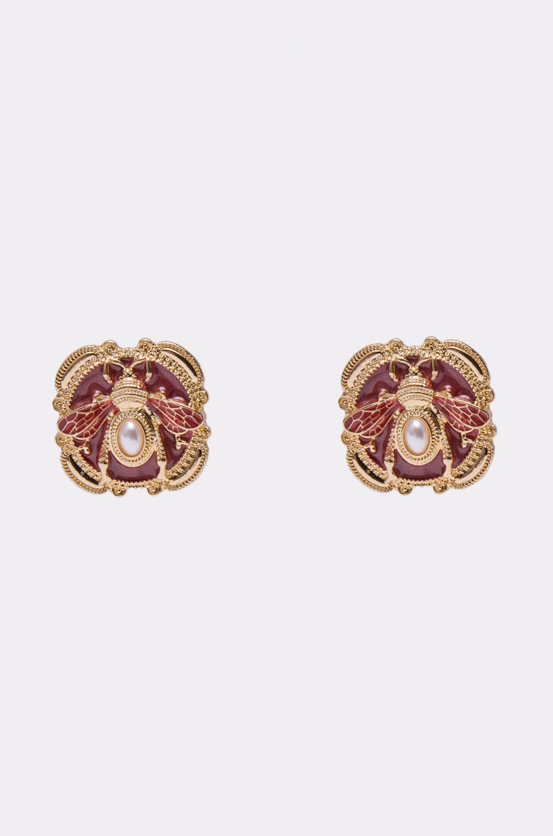 The DESI EARRING is a pair of ornate gold earrings with red accents and a central faux pearl, featuring intricate designs, displayed on a plain white background. 0