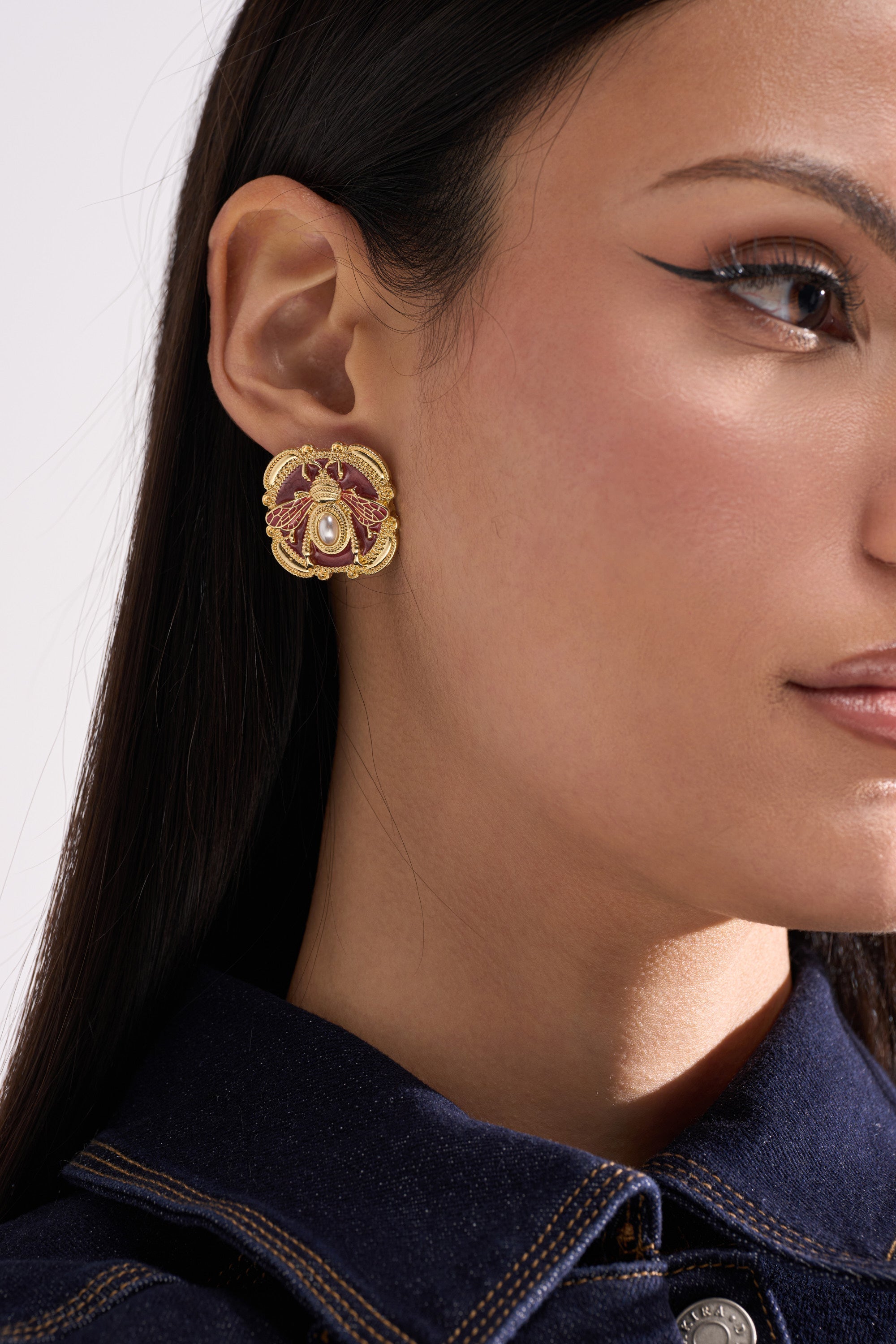 Close-up of a woman in a dark denim shirt and winged eyeliner wearing DESI EARRING—large, ornate gold earrings with red accents and a central faux pearl. 1