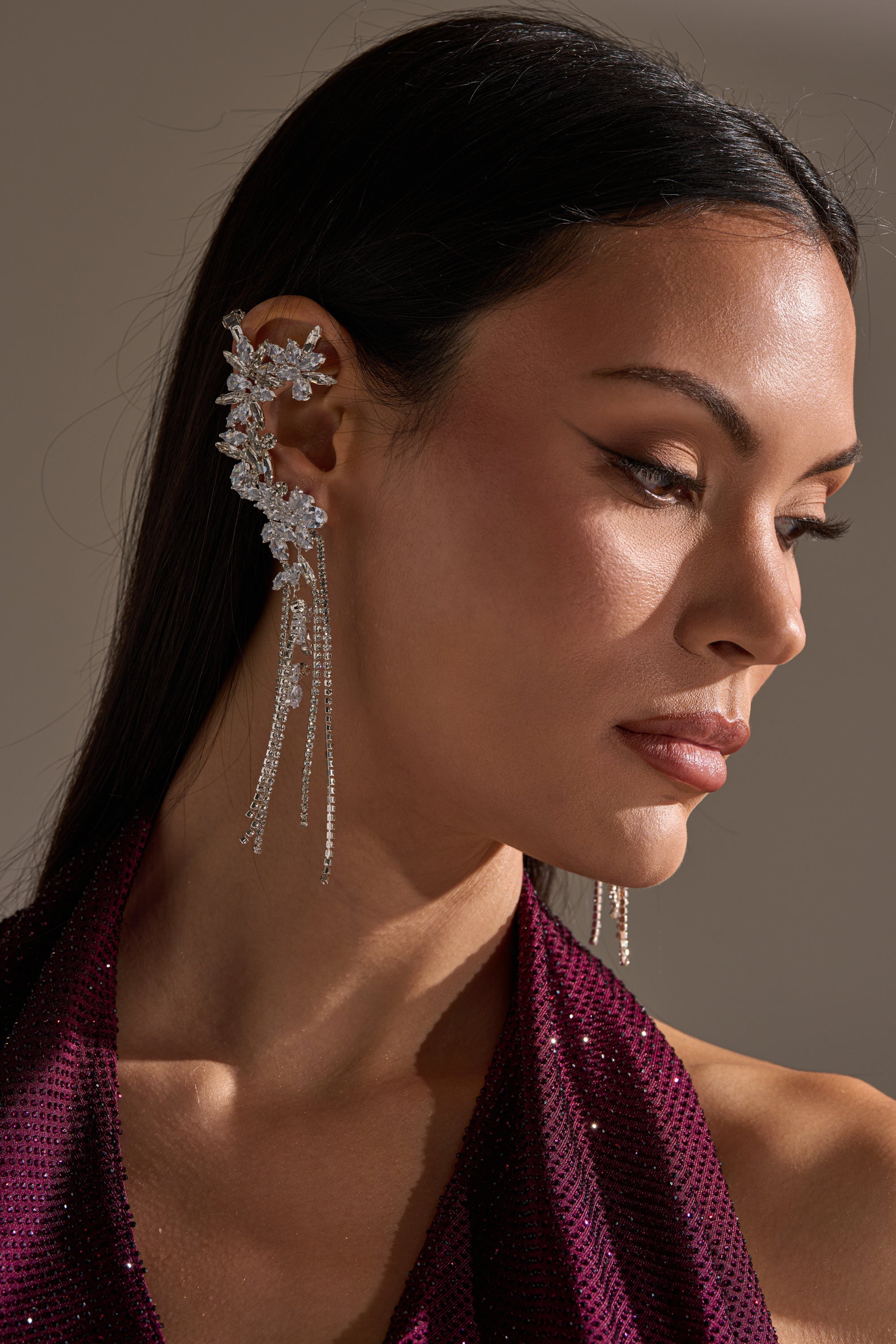 A woman with long, straight dark hair wears a maroon halter top and the KEL EARRING: an intricate silver ear cuff featuring crystal rhinestones, floral designs, and dangling chains. She gazes downward in soft lighting. 0