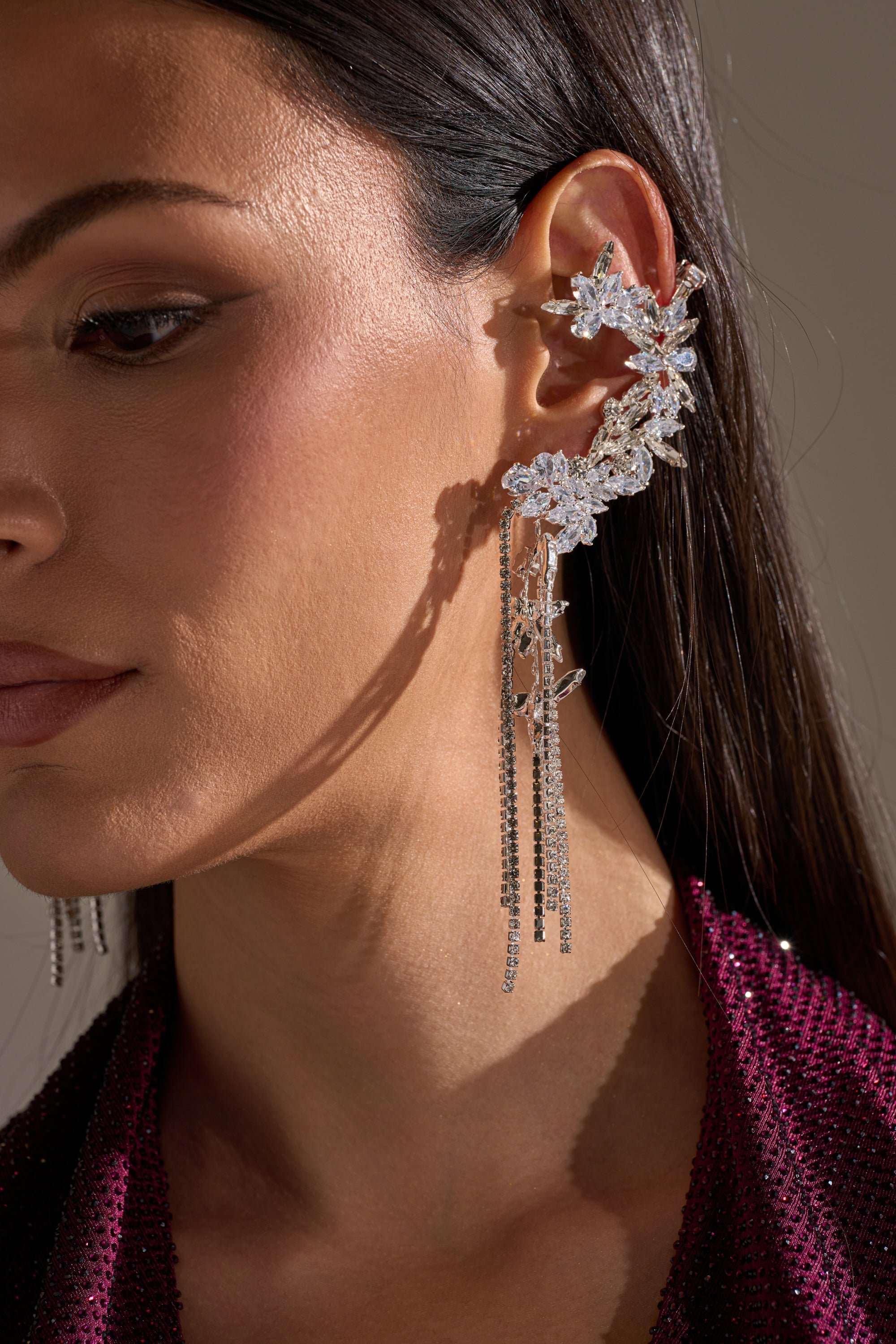 Close-up of a woman in straight dark hair and a maroon textured top, showcasing the KEL EARRING—an ornate ear cuff adorned with sparkling crystals and dangling silver chains. This statement piece adds instant glamour to her look. 2