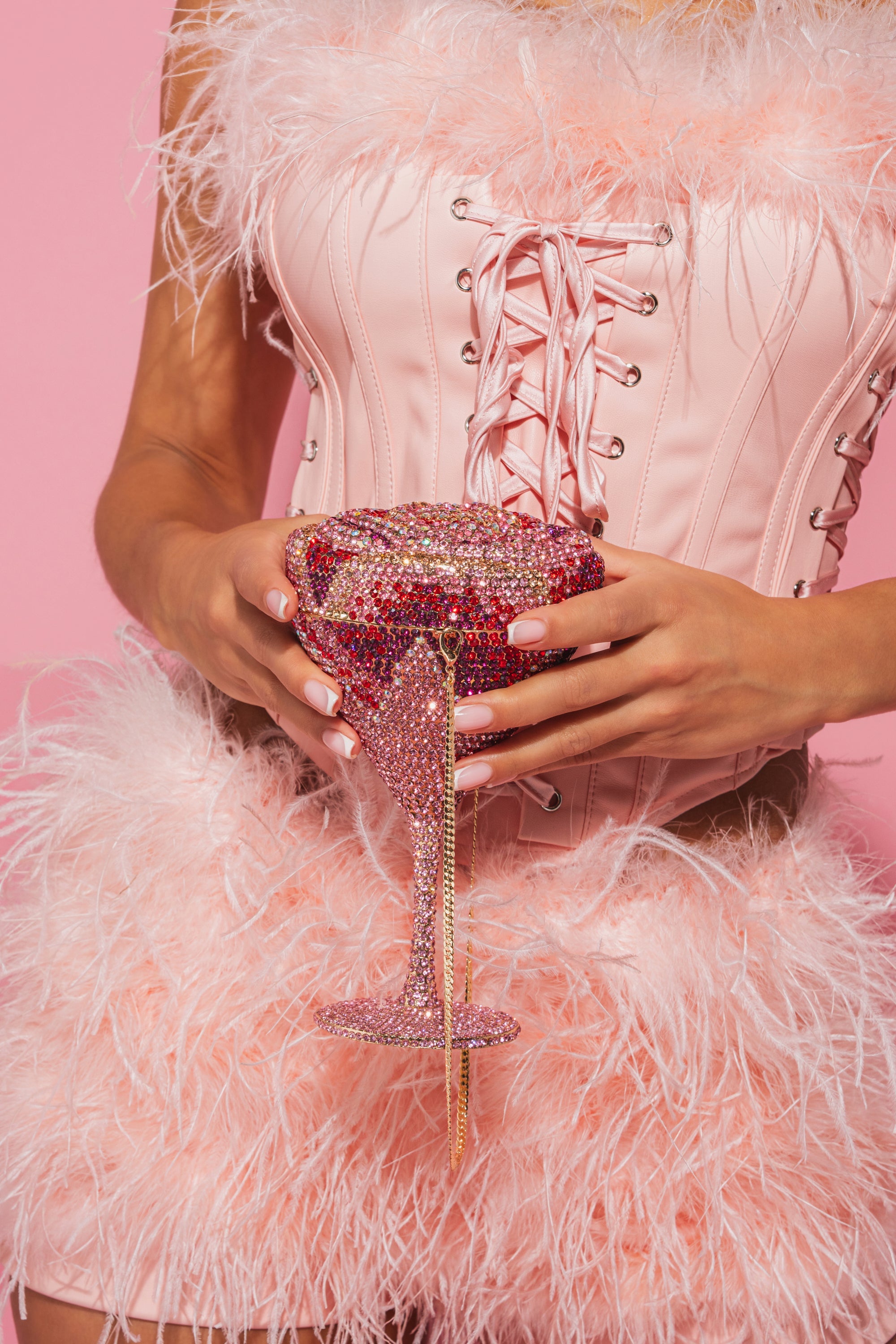 A person in a pink feathered outfit and corset holds the TAKE A SIP BLING PURSE, an ornate, rhinestone-encrusted statement accessory, against a pink background. 1