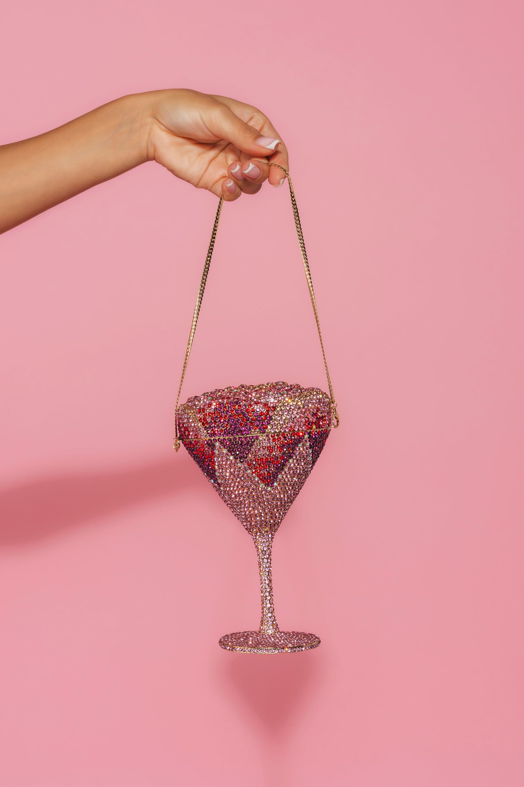 A manicured hand holds the TAKE A SIP BLING PURSE—a sparkling martini glass clutch with a thin gold chain—against a solid pink background, making it a true statement accessory. 0